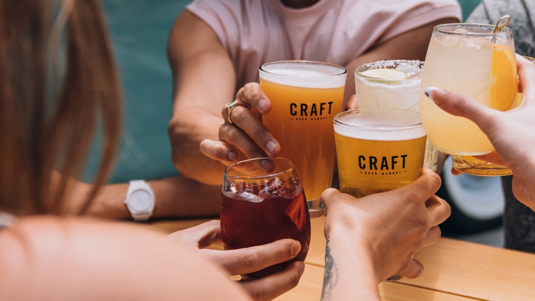 CRAFT Beer Market Five people clink glasses of craft beers and cocktails at a table; some glasses are branded with the word "CRAFT." Perfect for group bookings at multiple locations.
