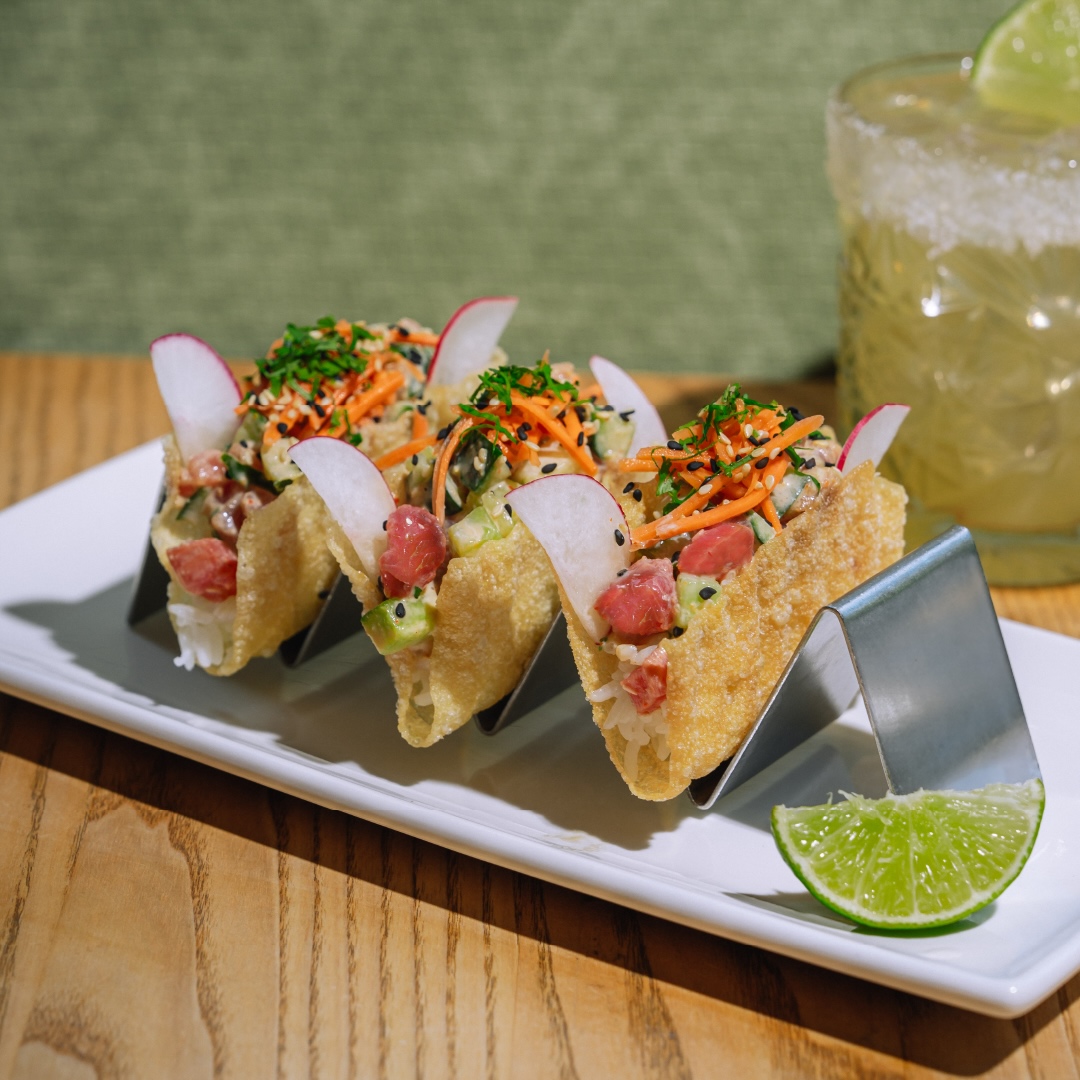 CRAFT Beer Market Three mini tacos filled with diced fish, vegetables, and garnishes are served on a metal taco holder with a lime wedge, next to a glass of craft cocktail with a salted rim and lime slice—perfect for casual dining.