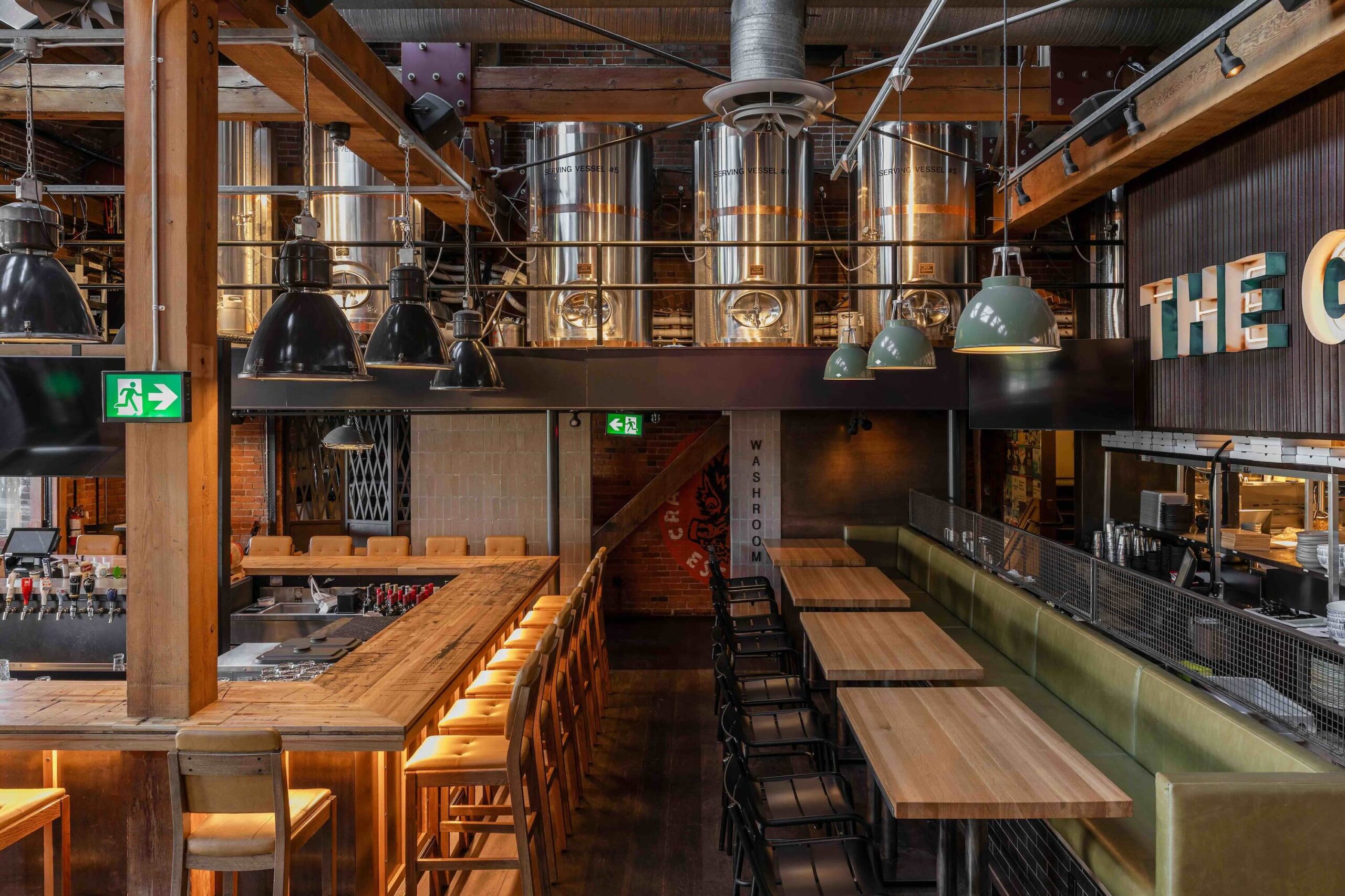CRAFT Beer Market Modern brewery interior featuring stainless steel tanks, a wooden bar with stools, high tables and chairs, green accent lighting, and industrial decor—perfect for enjoying craft beer and soaking in lively social vibes.