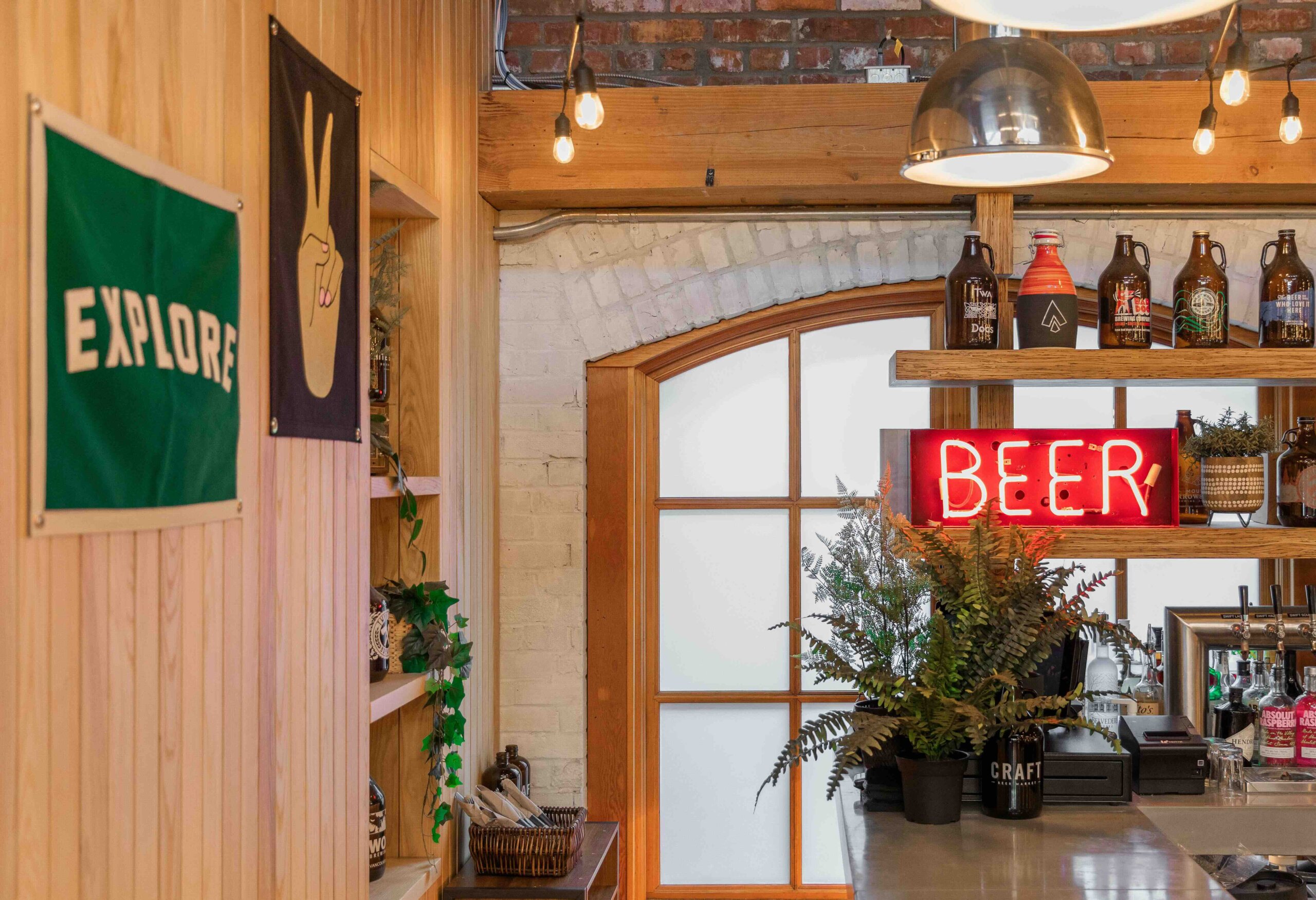 CRAFT Beer Market A bar area with a neon "BEER" sign, bottled drinks on shelves, hanging lights, plants, and a wooden wall with banners that read "EXPLORE" and a peace sign.