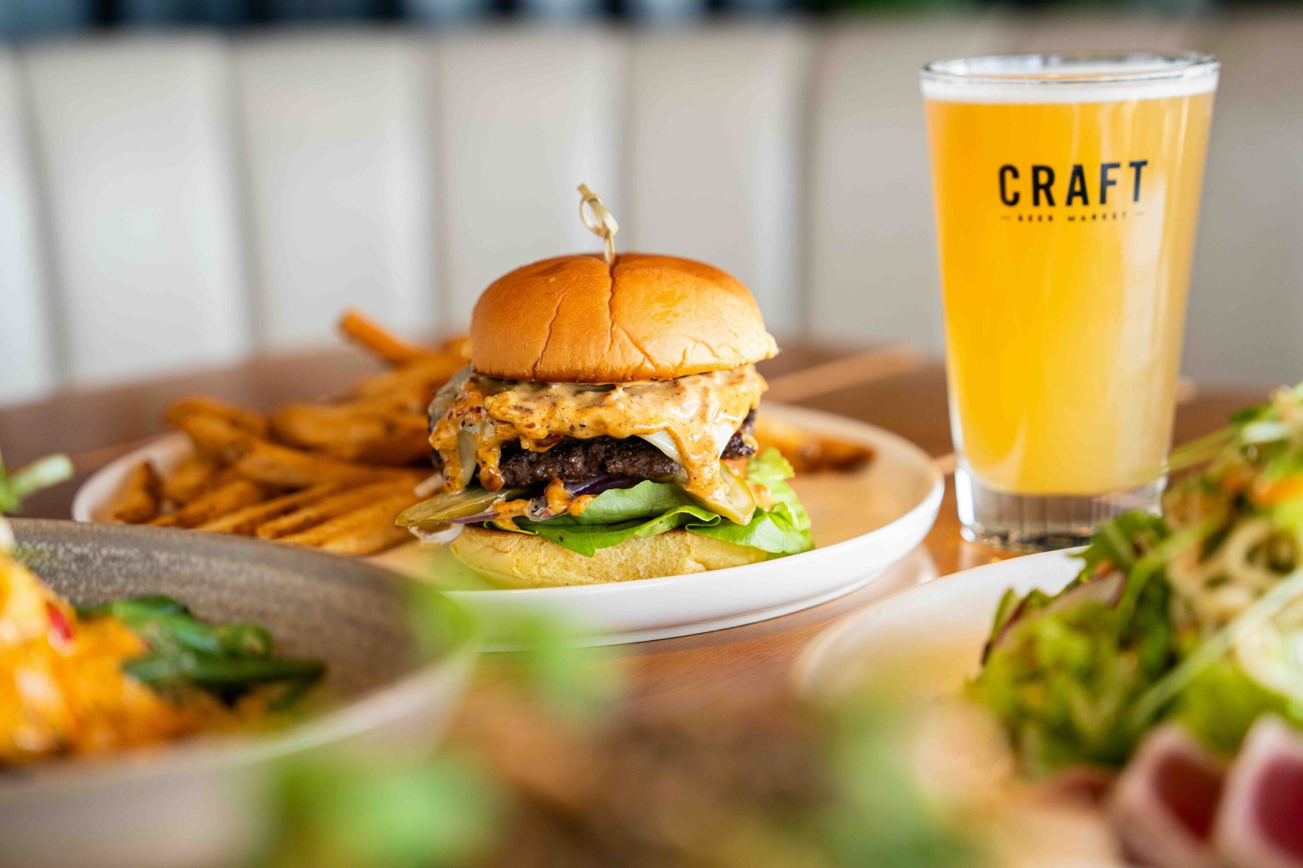 CRAFT Beer Market A cheeseburger with lettuce on a plate, served with fries and a glass of craft beer labeled "CRAFT," sets the perfect scene for unwinding and enjoying social vibes, with other dishes visible in the background.