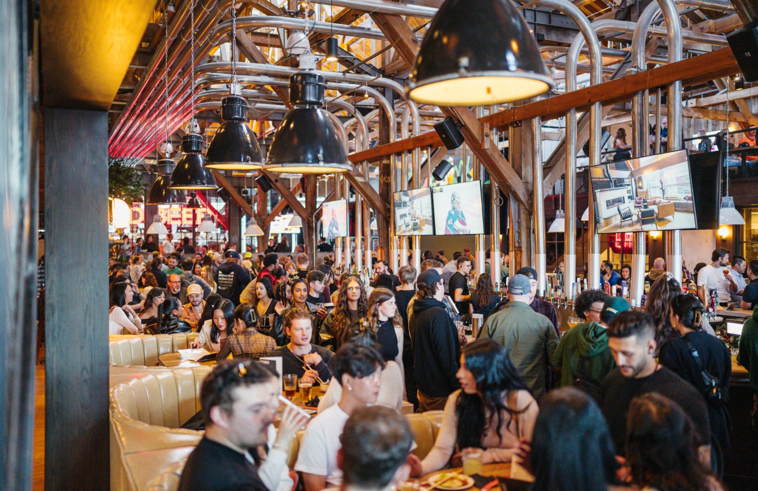 CRAFT Beer Market A large crowd of people socialize, eat, and drink inside a spacious, industrial-style restaurant with TVs and wooden beams overhead.