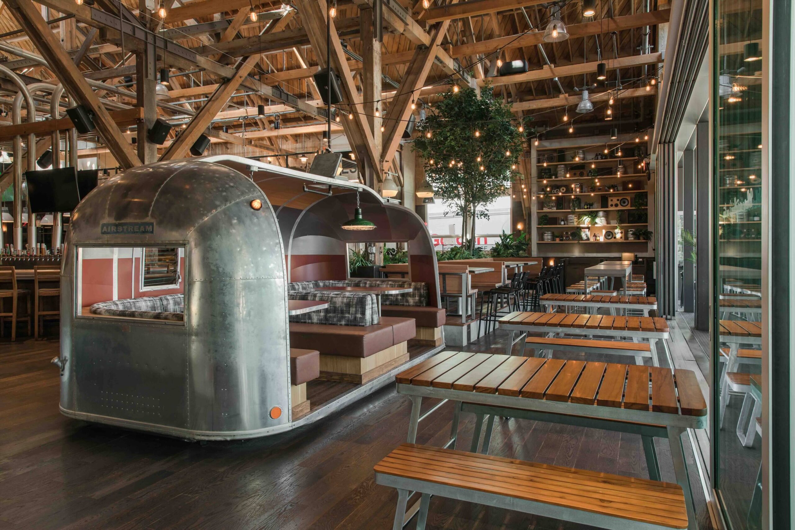 CRAFT Beer Market Indoor restaurant with wooden tables, an Airstream trailer booth, exposed beams, string lights, and plants by large windows—perfect for social vibes and enjoying craft cocktails.