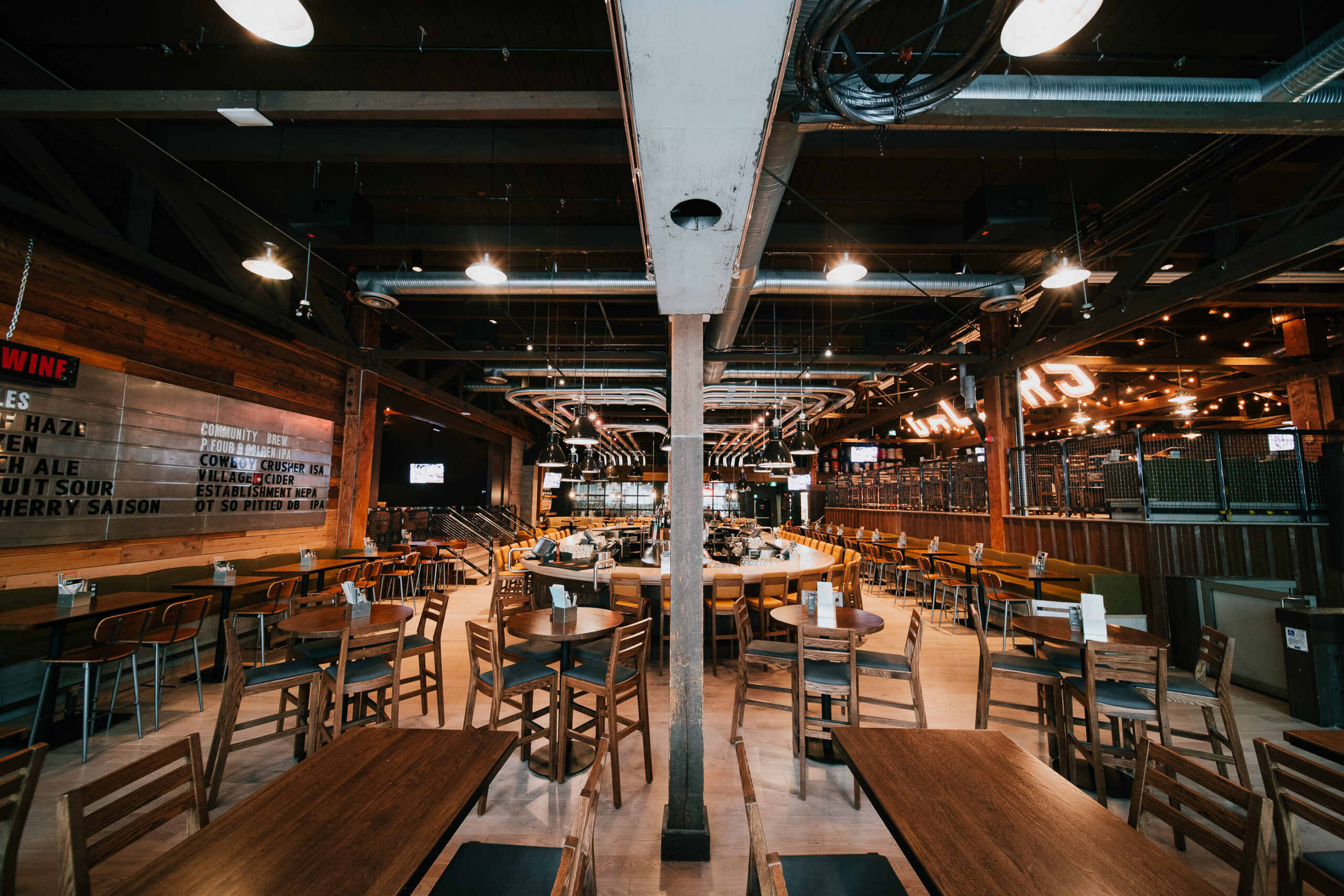 CRAFT Beer Market Spacious, modern restaurant with wooden tables and chairs, high ceilings, and industrial lighting, viewed from the center aisle toward the bar area. Perfect for group bookings and featuring a selection of craft beers.