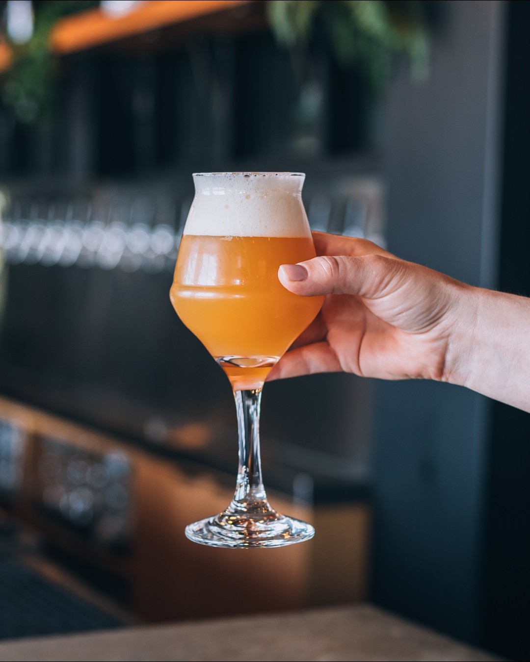 CRAFT Beer Market A hand holds a stemmed glass filled with hazy, golden beer topped with a frothy head in a bar setting, perfect for group bookings across multiple locations.
