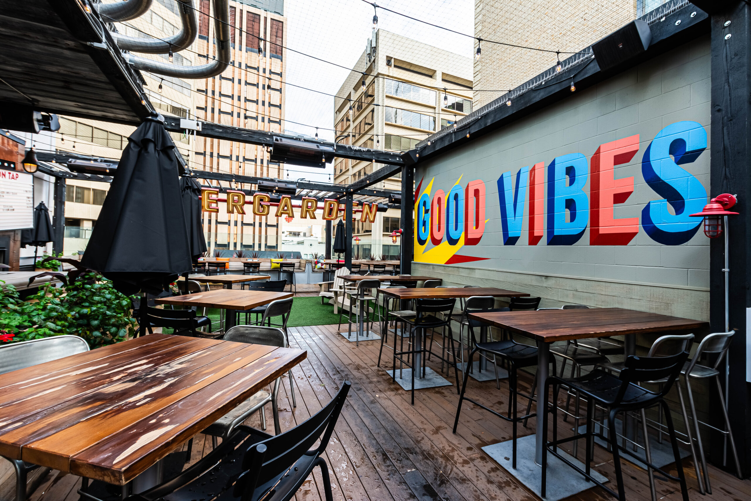 CRAFT Beer Market Outdoor rooftop bar area with empty wooden tables and chairs, surrounded by buildings; a mural reads "GOOD VIBES" on one wall. Enjoy sustainable craft cocktails in this inviting urban setting.