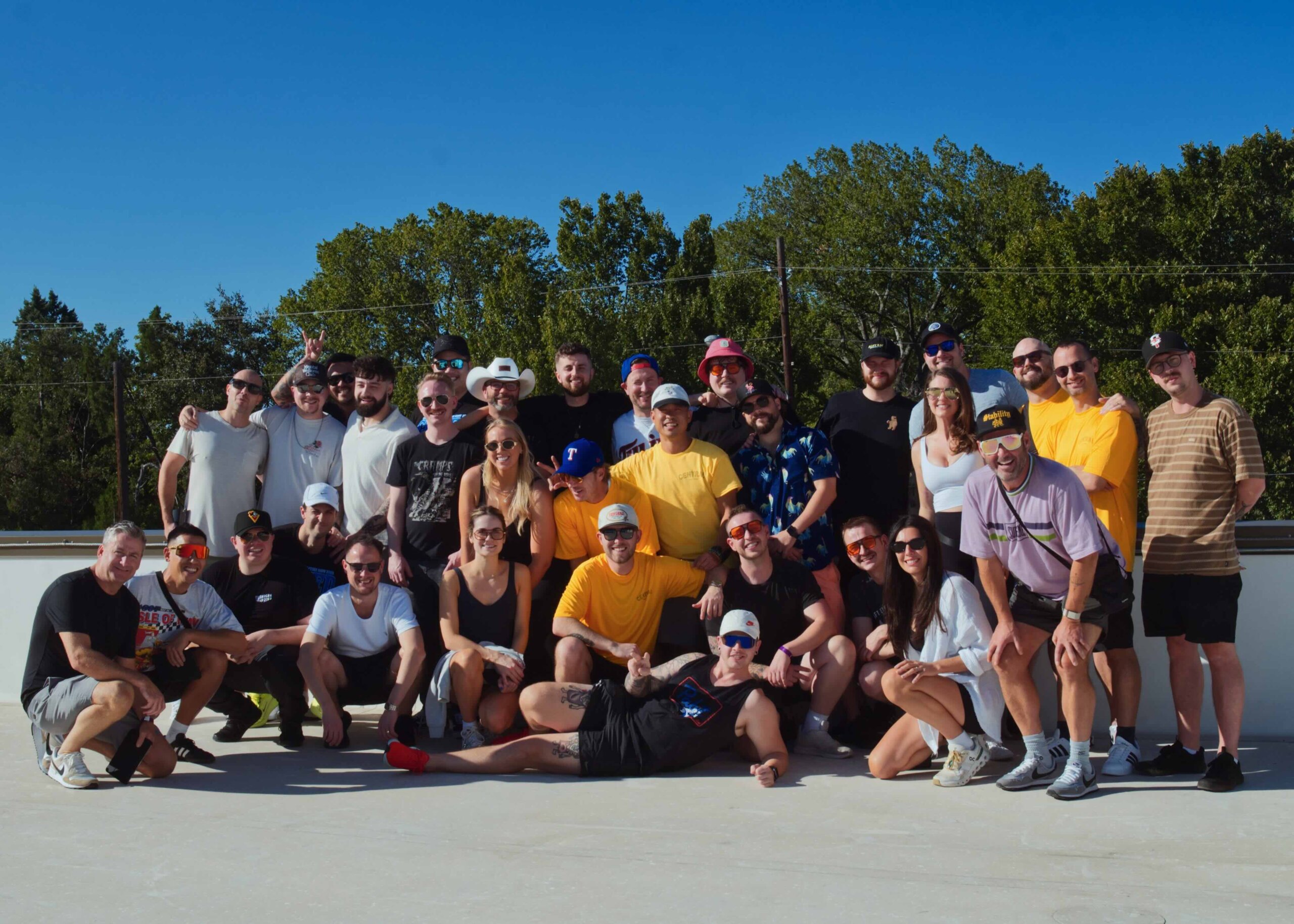 CRAFT Beer Market A group of people pose together outdoors on a sunny day, standing and kneeling on a concrete surface with trees and a blue sky in the background.