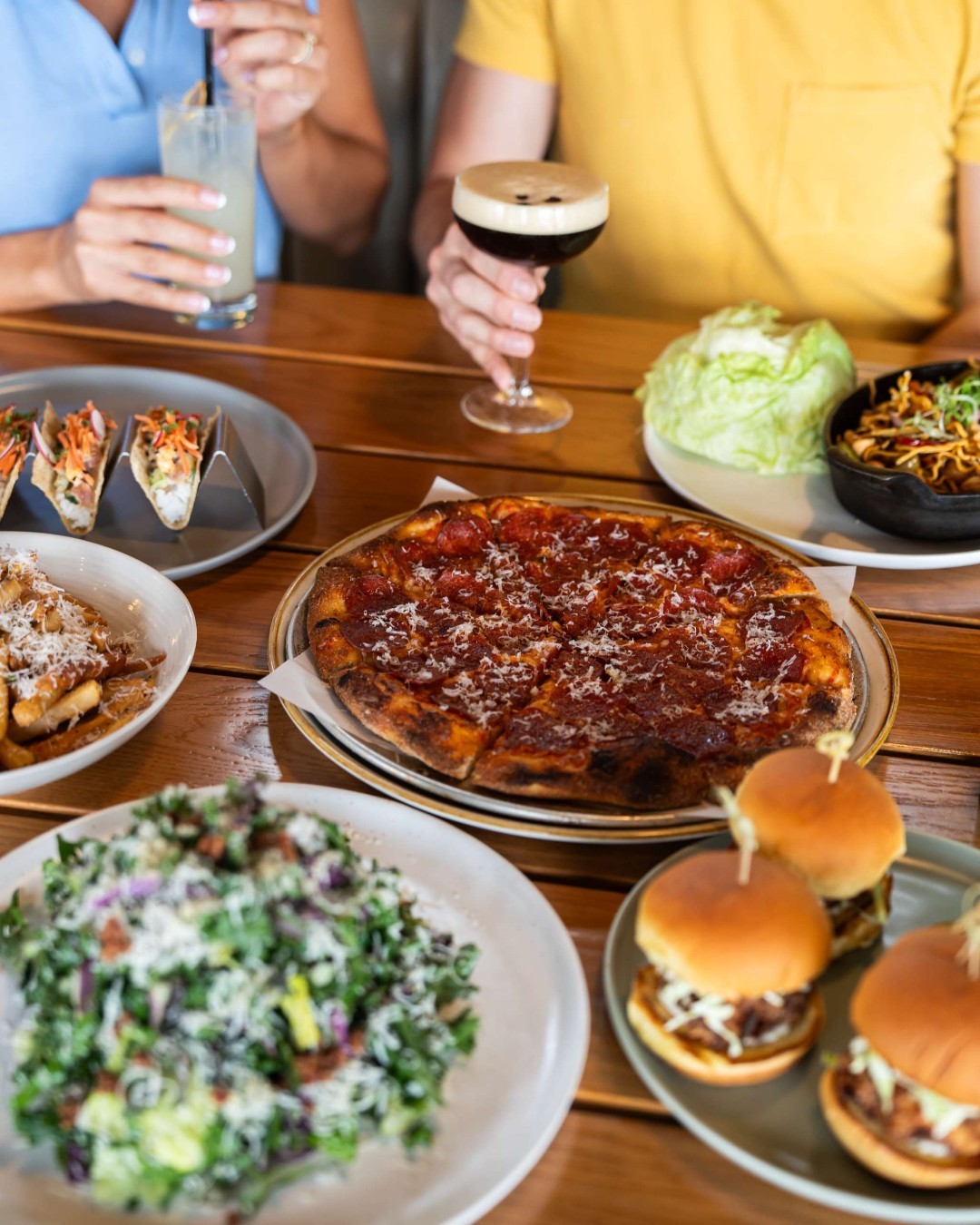 CRAFT Beer Market A table with pizza, salad, sliders, tacos, and drinks, with two people enjoying craft cocktails in the background.