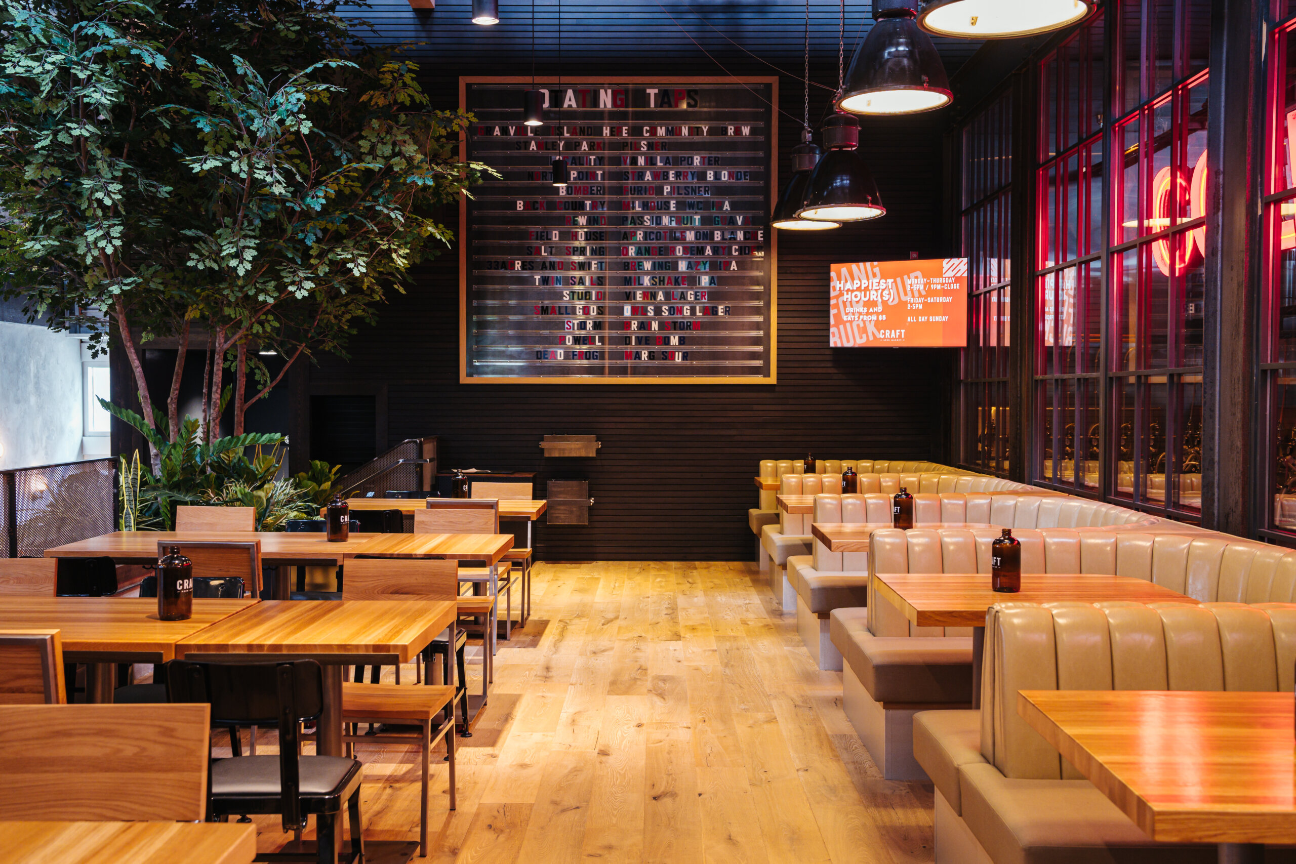 CRAFT Beer Market A modern restaurant interior with wooden tables, tan upholstered booths, chairs, indoor plants, and a large wall menu board. The space is well-lit with hanging lights and filled with social vibes—perfect for enjoying craft cocktails.
