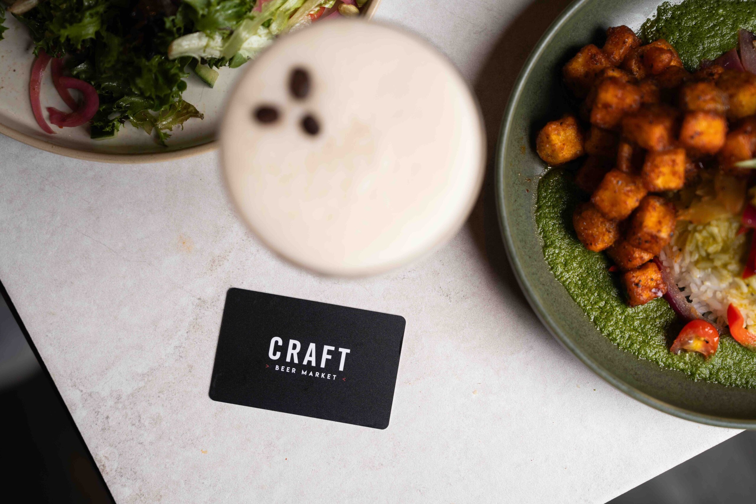 CRAFT Beer Market A black "Craft Beer Market" card on a white table next to a frothy drink and a plate of food with seasoned cubes and greens.
