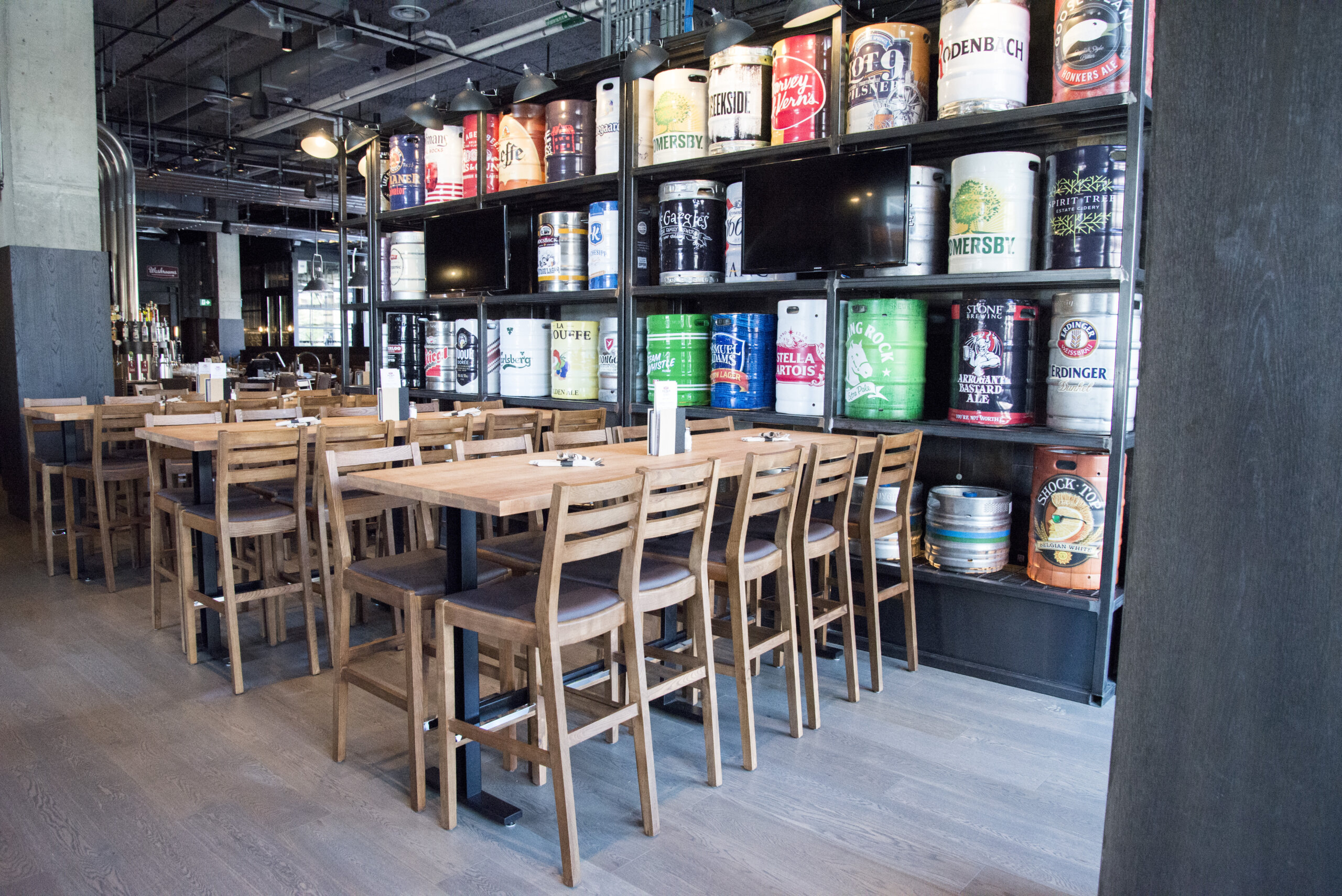 CRAFT Beer Market A modern bar with wooden tables and chairs, shelves displaying large colorful beer kegs, and an impressive menu of craft cocktails—perfect for group bookings.