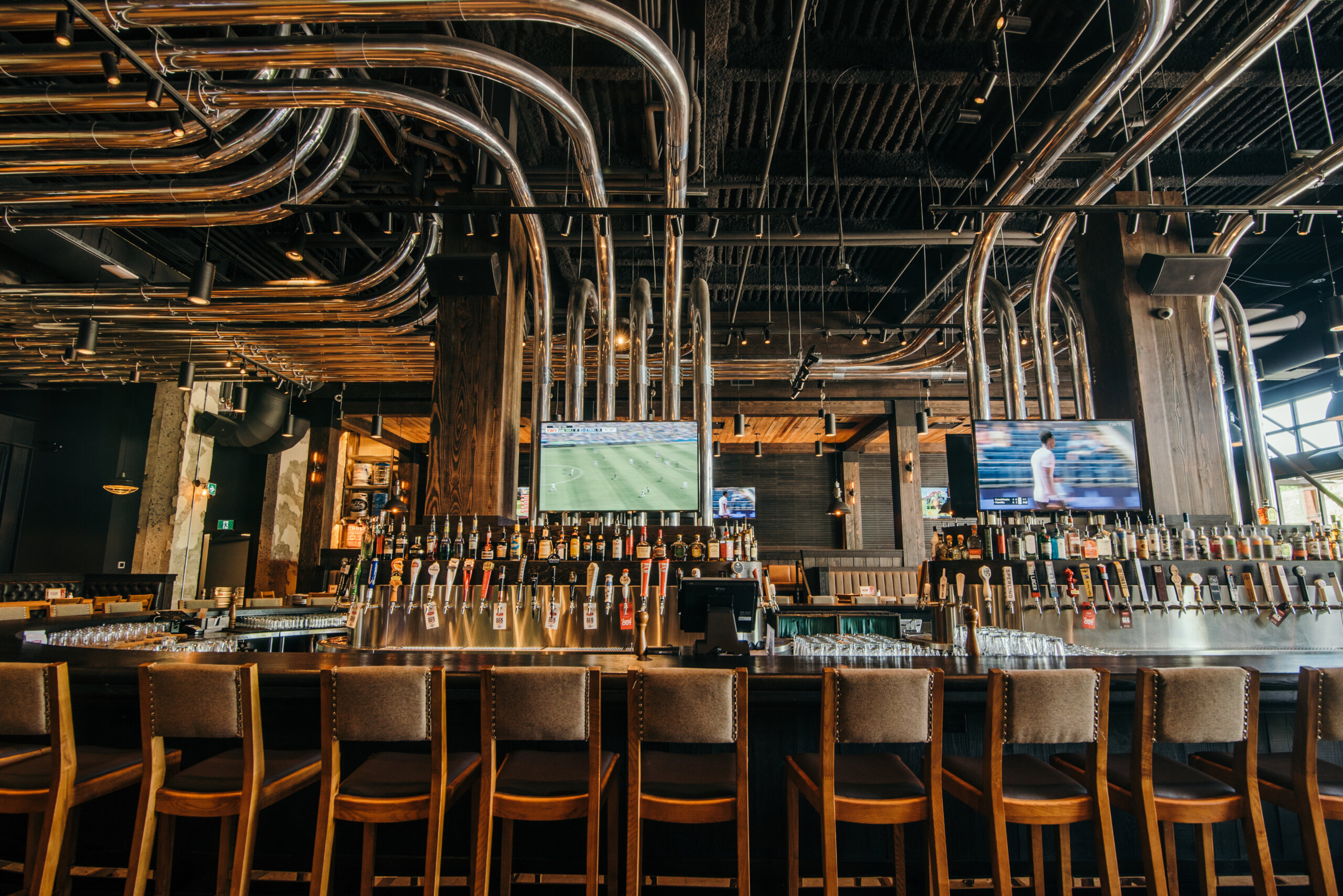CRAFT Beer Market Modern bar with a long counter, rows of rotational taps, wooden chairs, metallic pipes on the ceiling, and two TVs displaying sports—perfect for enjoying craft cocktails and social vibes.
