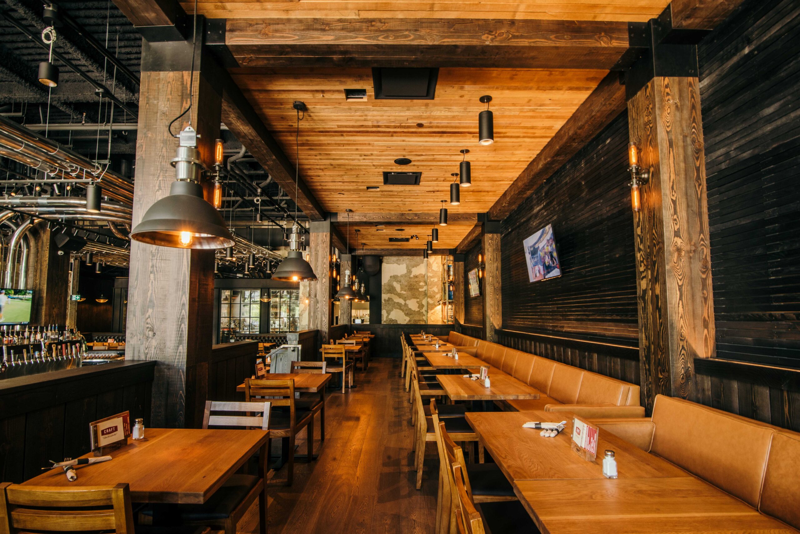 CRAFT Beer Market A modern restaurant interior with wooden tables and chairs, brown leather seating, hanging industrial lights, and a wall-mounted TV. Enjoy social vibes in this well-lit space, perfect for sharing craft beer with friends.
