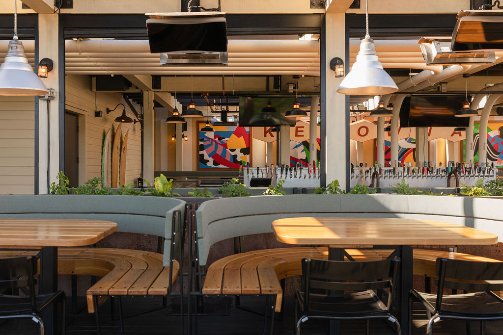 CRAFT Beer Market Outdoor dining area with wooden curved benches and black chairs, facing an indoor bar with vibrant wall art, rotational taps, and multiple ceiling-mounted TVs—perfect for enjoying craft cocktails and lively social vibes.