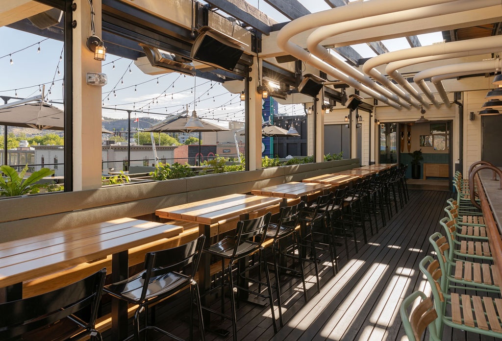 CRAFT Beer Market Outdoor rooftop dining area with long wooden tables, black chairs, string lights, potted plants, and large pipes on the ceiling, with views of nearby buildings and hills.