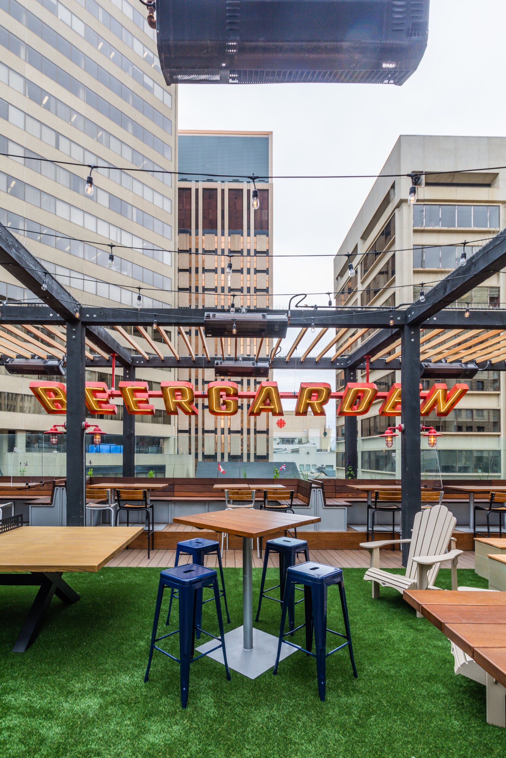 CRAFT Beer Market Outdoor rooftop beer garden with artificial grass, wooden tables, barstools, craft beer on rotational taps, and a large neon "Beergarden" sign, surrounded by tall office buildings.