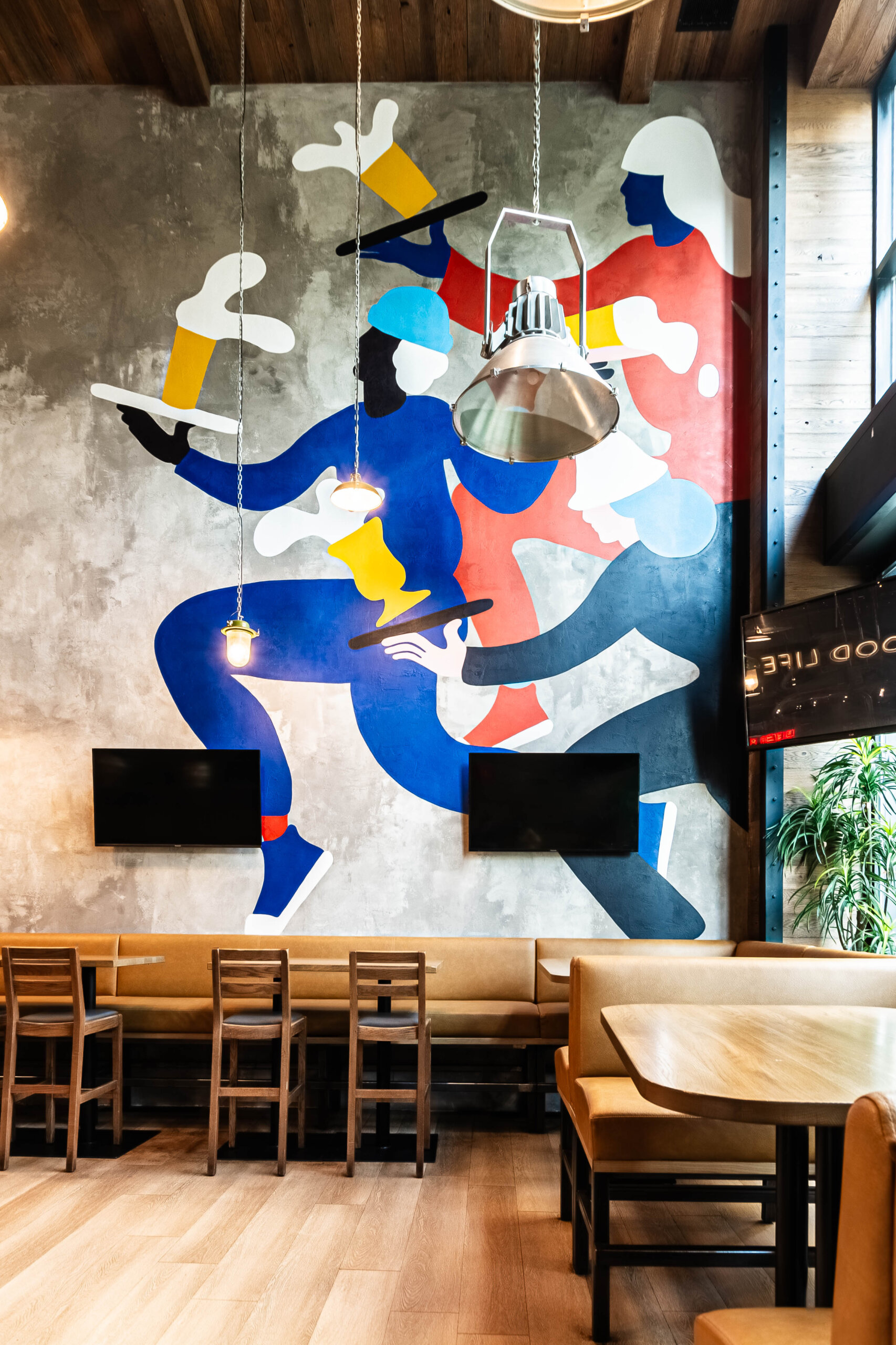 CRAFT Beer Market A modern restaurant interior with sustainable wooden tables and chairs, a large mural of abstract servers carrying food and drinks on the wall, and two flat-screen TVs below the mural.