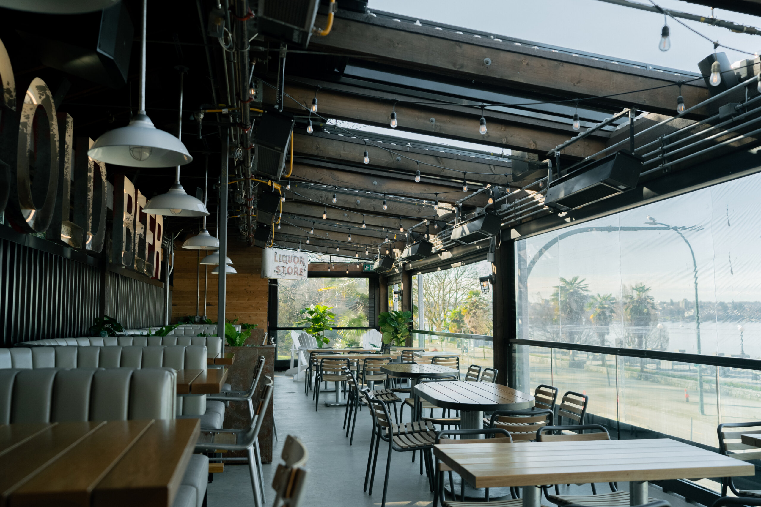 CRAFT Beer Market Modern restaurant with wooden tables and chairs, hanging lights, large windows, and a view of trees and water. The space is empty, filled with natural light—perfect for private events or enjoying craft cocktails in a serene setting.