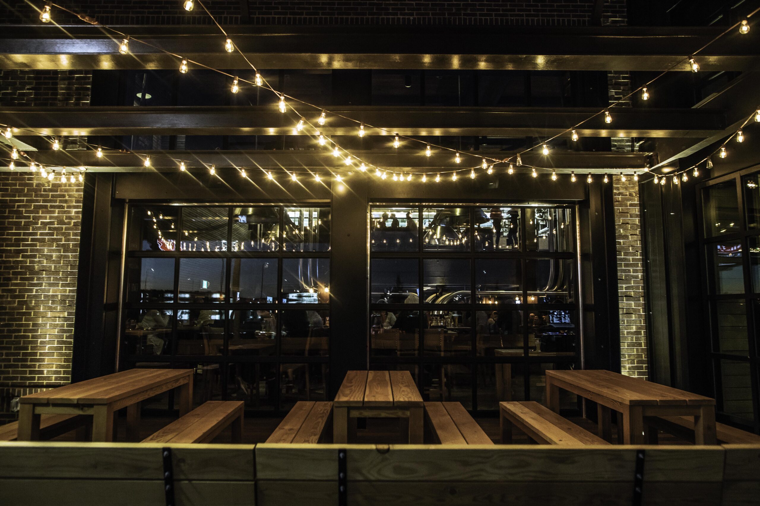 CRAFT Beer Market Outdoor patio with wooden picnic tables, string lights overhead, and large windows reflecting indoor lights in a brick-walled setting at night—perfect for enjoying craft beer and social vibes.
