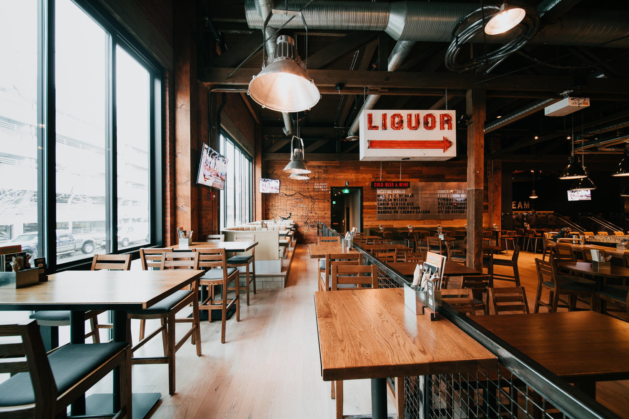 CRAFT Beer Market Spacious restaurant interior with wooden tables and chairs, industrial lighting, large windows, and a ceiling sign reading "LIQUOR" with an arrow pointing right—perfect for social vibes and enjoying a craft beer.