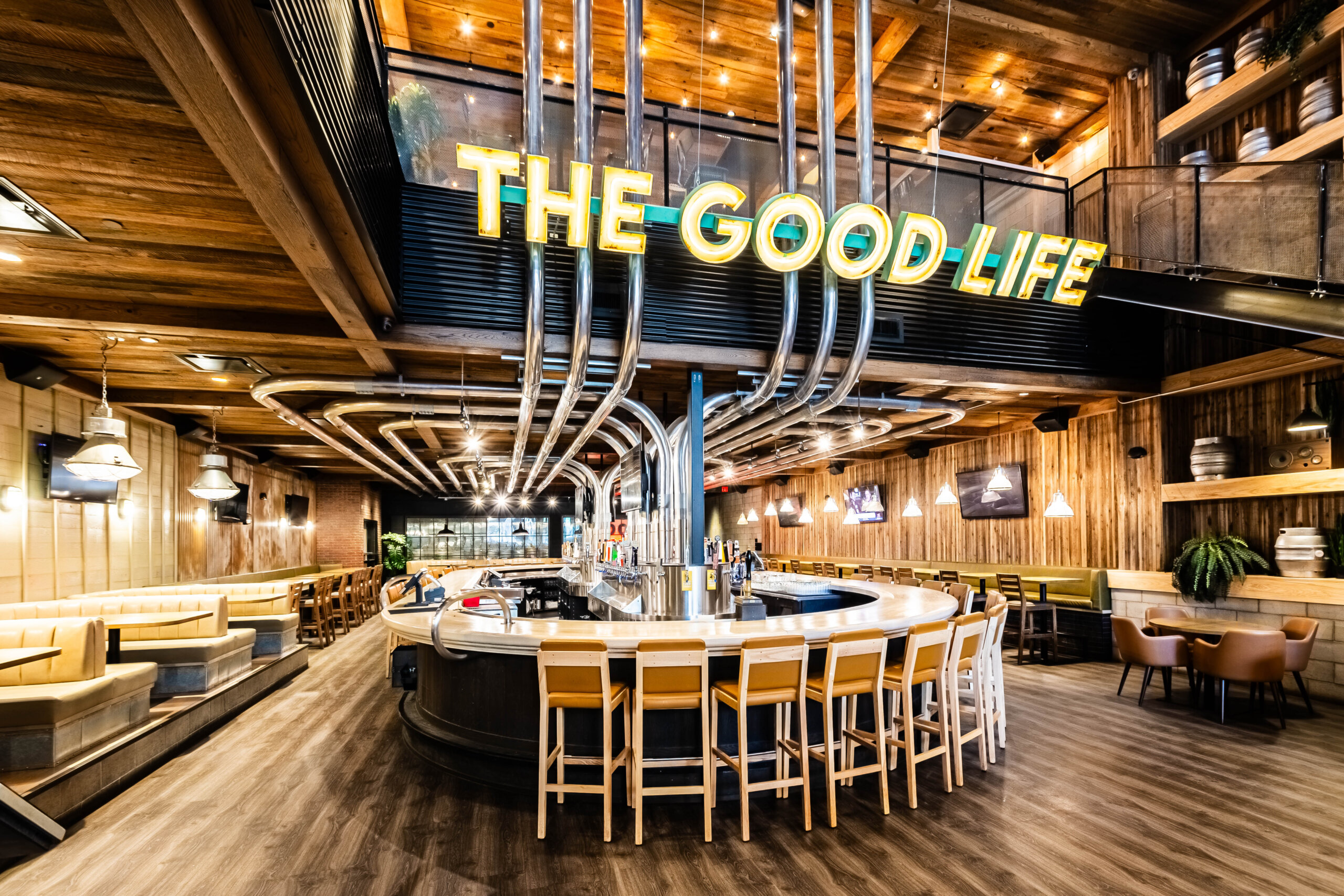 CRAFT Beer Market Modern bar with curved white counter, yellow chairs, wood paneling, and a large neon sign reading "THE GOOD LIFE" overhead. Enjoy craft beer from rotational taps beneath exposed pipes and warm lighting.