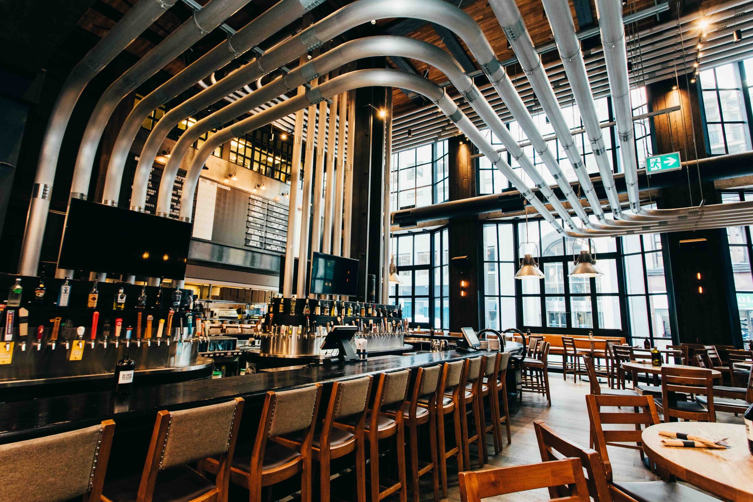 CRAFT Beer Market Modern bar interior with high ceilings, large windows, multiple beer taps, wooden chairs, and curved metal pipes running across the ceiling.