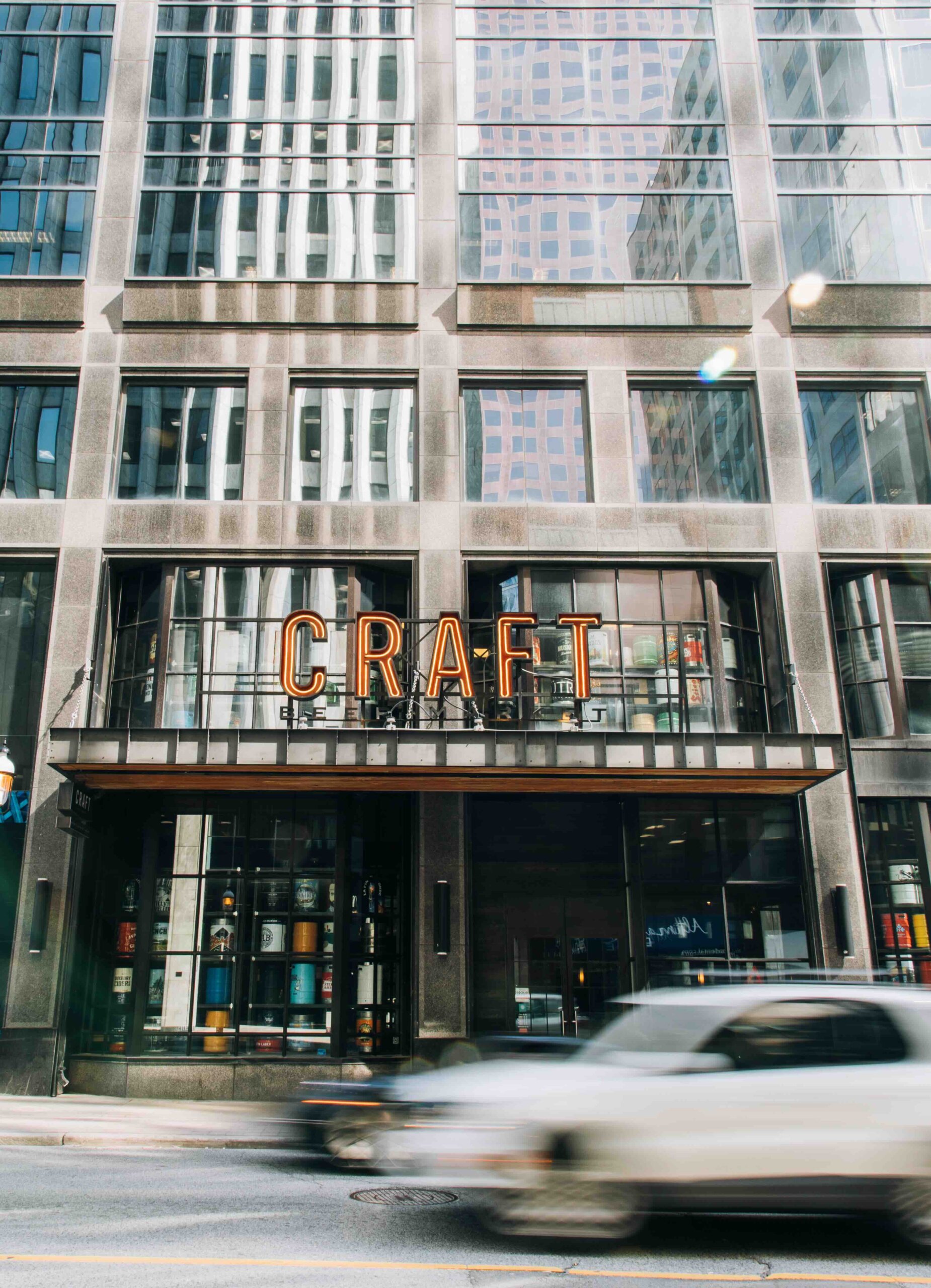 CRAFT Beer Market Street view of a glass and steel building with a large "CRAFT" sign above the entrance; a blurred white car passes in the foreground.