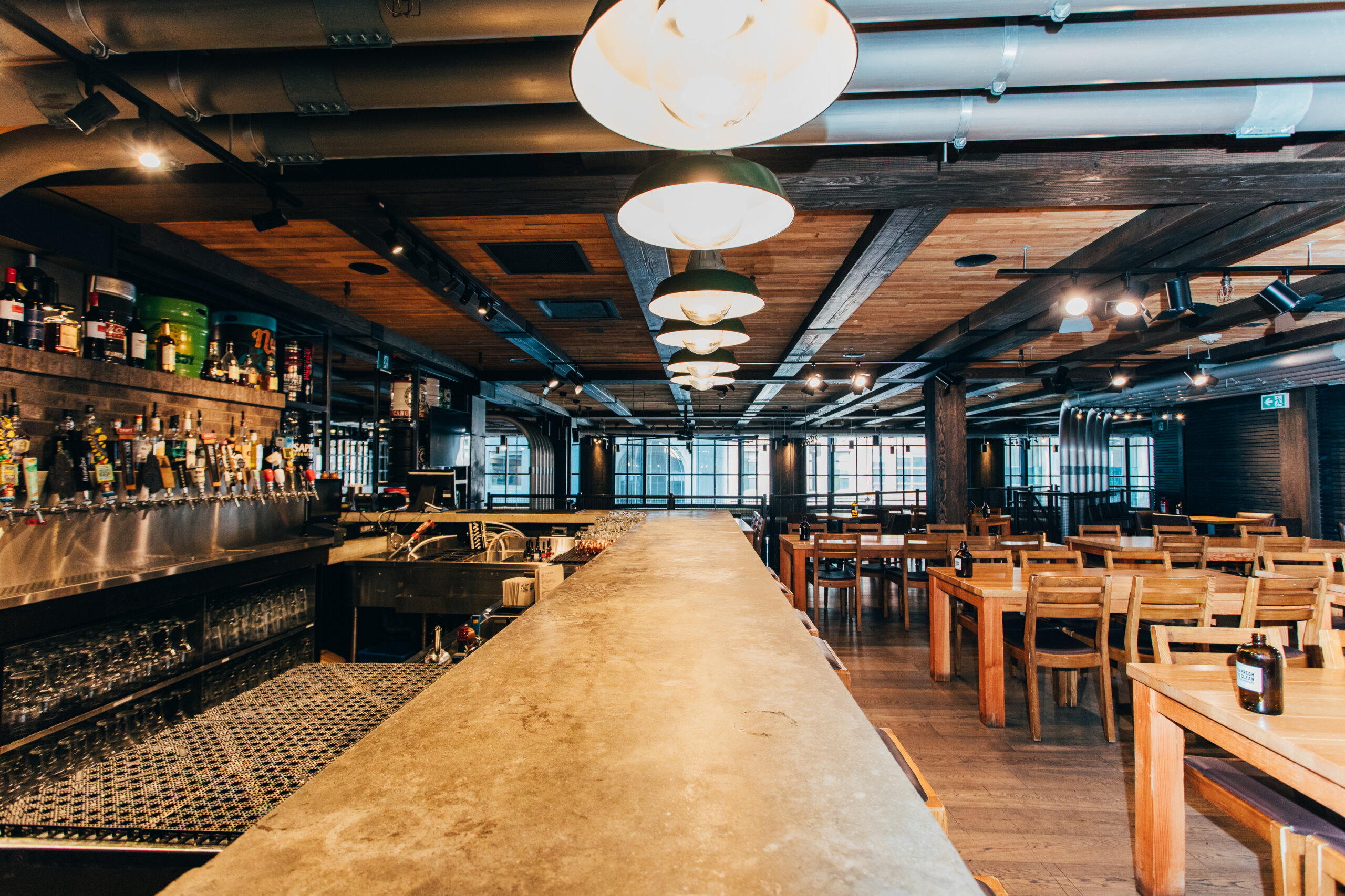 CRAFT Beer Market A spacious, modern bar with an empty countertop, shelves of liquor bottles, wooden tables and chairs, industrial-style ceiling lighting, and a menu featuring craft cocktails made with locally sourced ingredients.