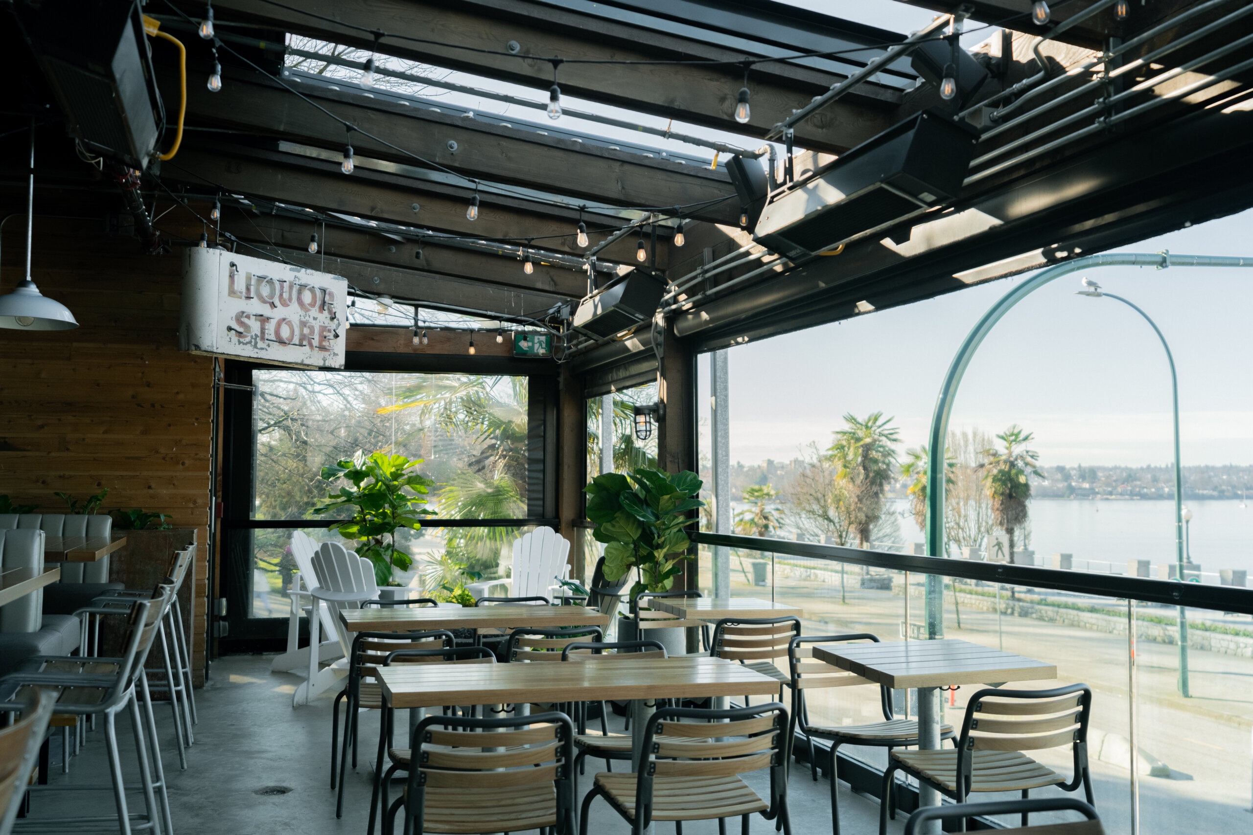 CRAFT Beer Market A covered outdoor patio with wooden tables and chairs, potted plants, string lights, and a "Liquor Store" sign, overlooking a waterfront view—perfect for sipping craft cocktails in a relaxed setting.