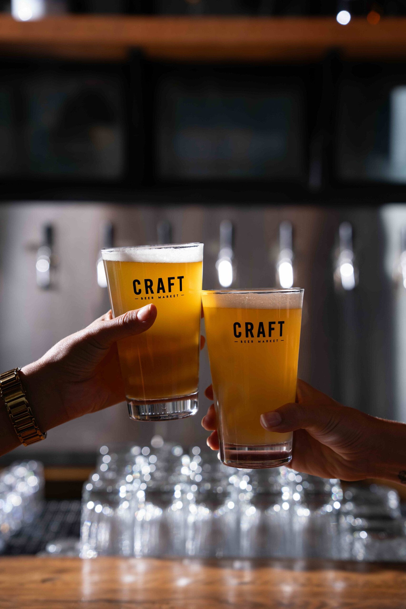 CRAFT Beer Market Two hands holding up pints of beer labeled "CRAFT" in a bar setting, with beer taps and glasses visible in the background.