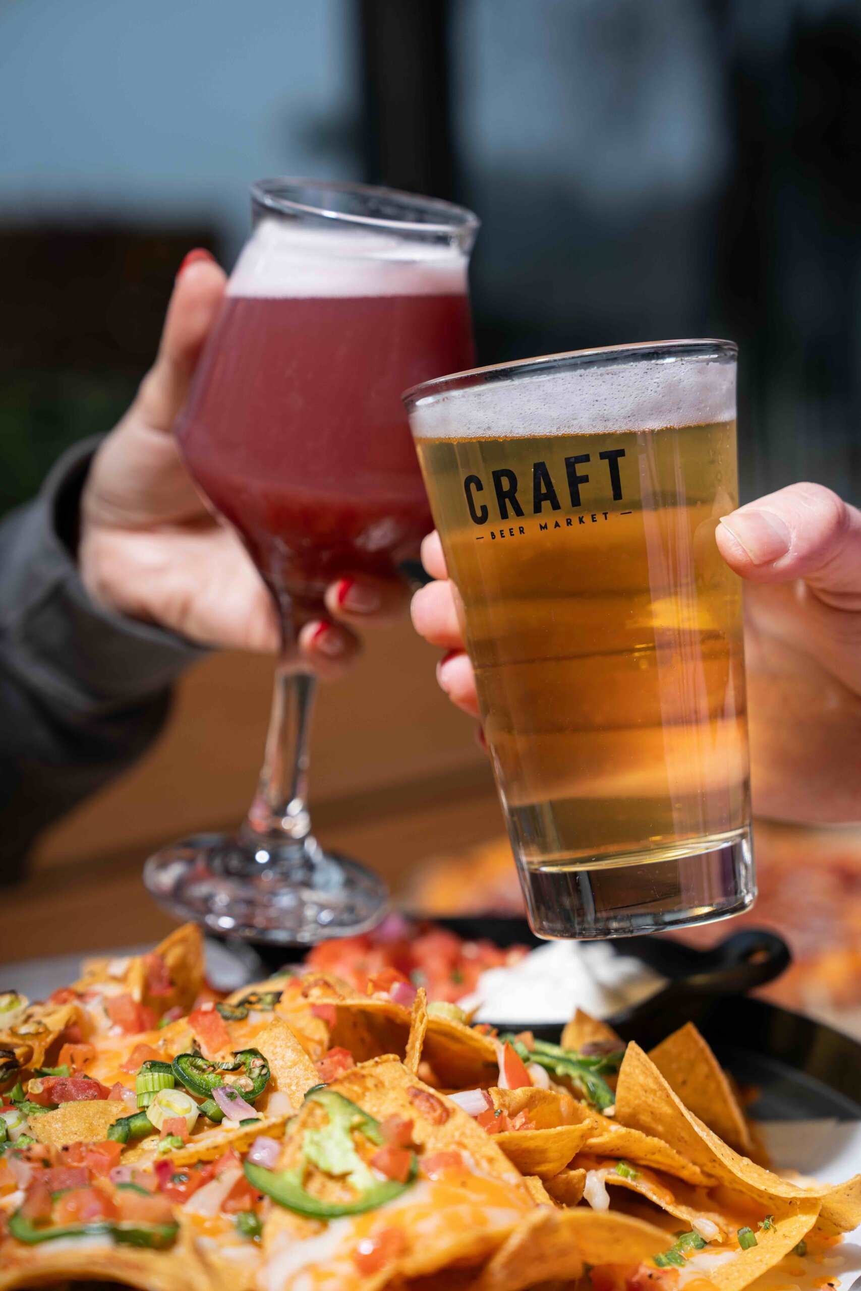 CRAFT Beer Market Two people clink glasses, one with a red drink and one with beer, over a plate of nachos topped with tomatoes, jalapeños, and cheese.