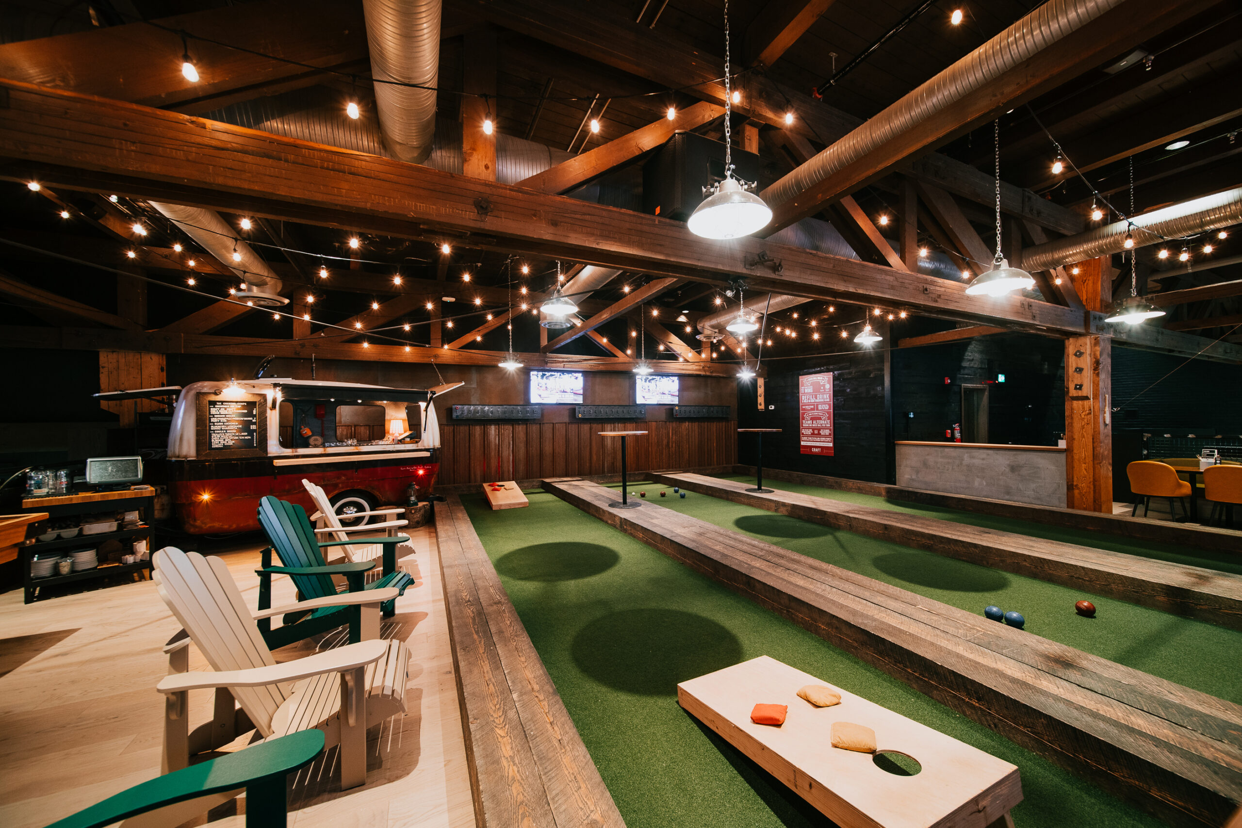 CRAFT Beer Market Indoor bocce ball court with turf lanes, wooden beams, string lights, Adirondack chairs, and a retro food truck; TVs, seating area, and a menu featuring craft cocktails are visible in the background.