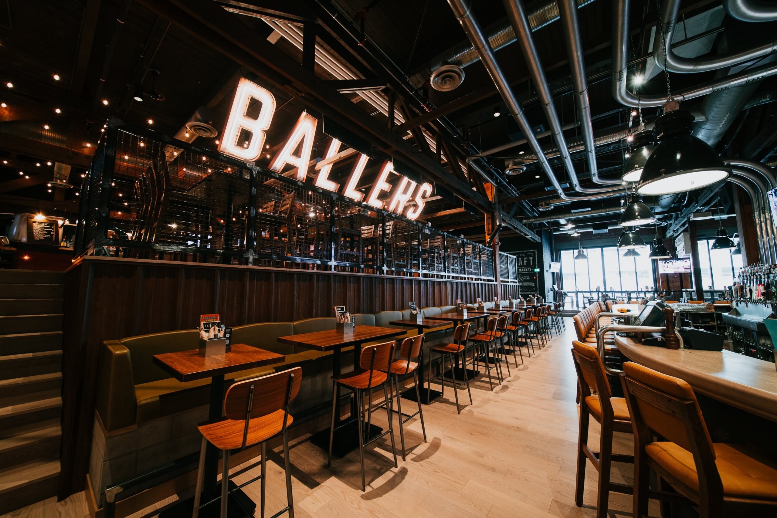 CRAFT Beer Market Modern restaurant interior with wooden tables and chairs, booth seating, and a large illuminated "BALLERS" sign above a glass-walled area.