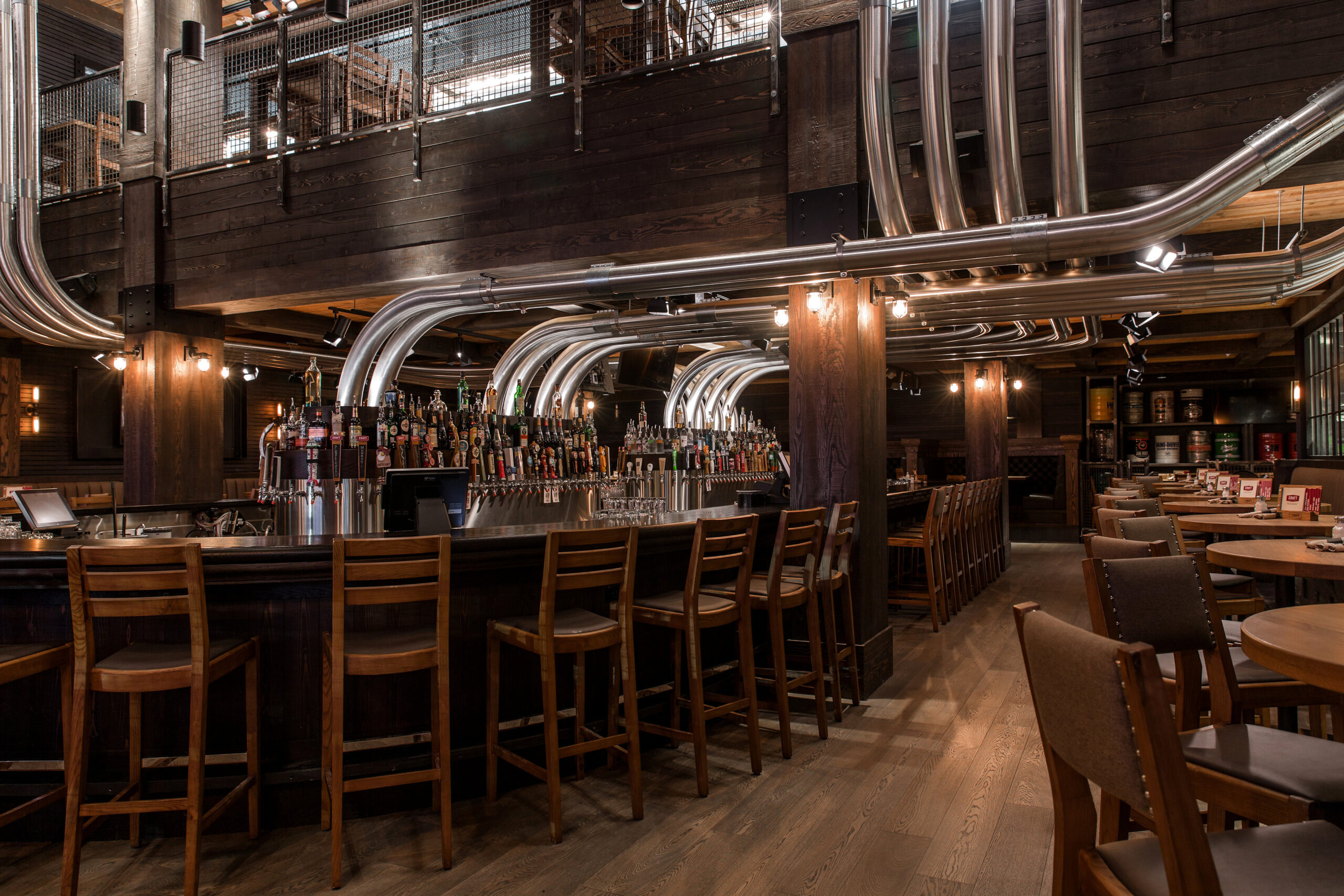CRAFT Beer Market A modern bar with wooden chairs and tables, metal pipe fixtures overhead, craft cocktails, and a variety of bottled and draft drinks displayed behind the counter. Perfect for group bookings.