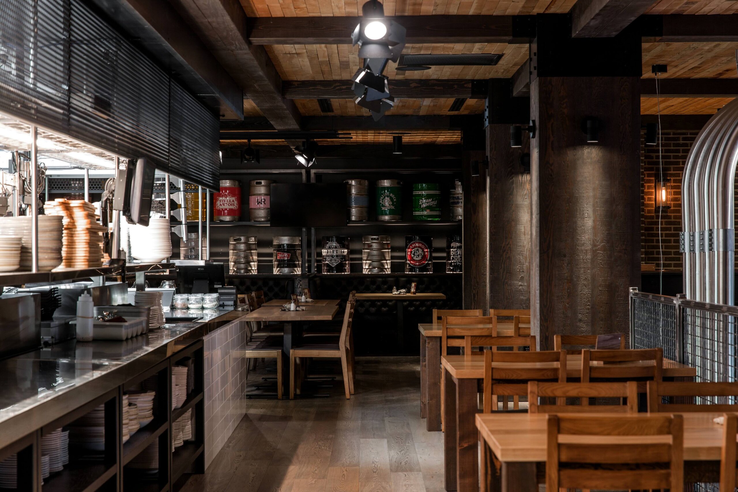CRAFT Beer Market A modern restaurant interior features wooden tables and chairs, a stainless steel counter with stacked plates, and shelves displaying various beer kegs in the background.