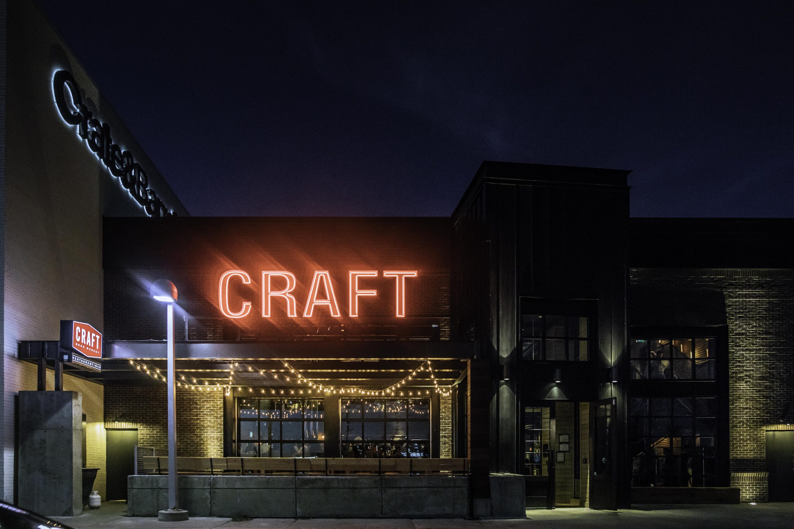 CRAFT Beer Market A restaurant exterior at night with a large red neon "CRAFT" sign above the entrance, string lights along the patio, and a dark brick facade—serving up craft cocktails in style.