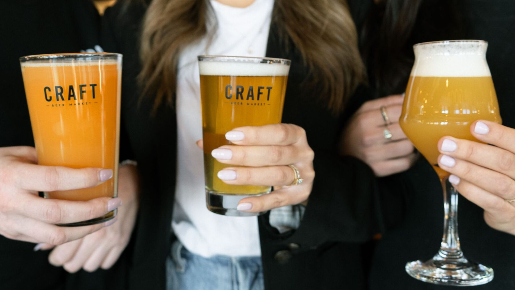 CRAFT Beer Market Three people holding craft beers in branded "CRAFT" glasses, each with light-colored nail polish and casual clothing visible—celebrating unique flavors enjoyed across multiple locations.