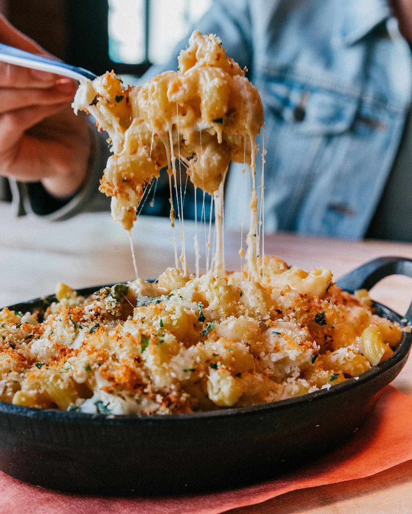 CRAFT Beer Market A person lifting a forkful of cheesy baked macaroni and cheese from a cast iron dish, with melted cheese stretching—perfect comfort food to enjoy with friends during group bookings at our multiple locations.