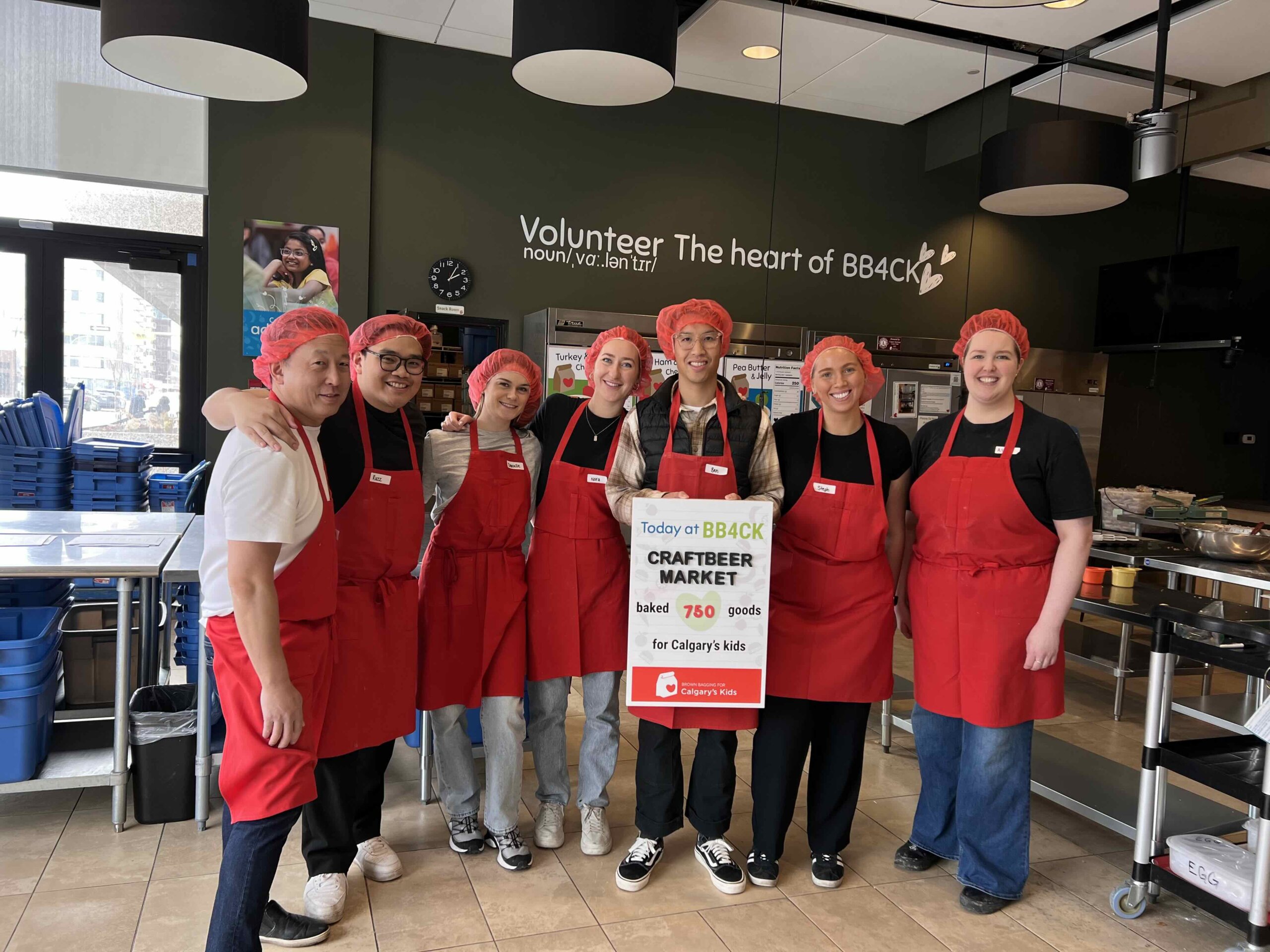 CRAFT Beer Market A group of eight people in red aprons and hairnets stand in a kitchen holding a sign about a bakery market for Calgary kids at BB4CK.
