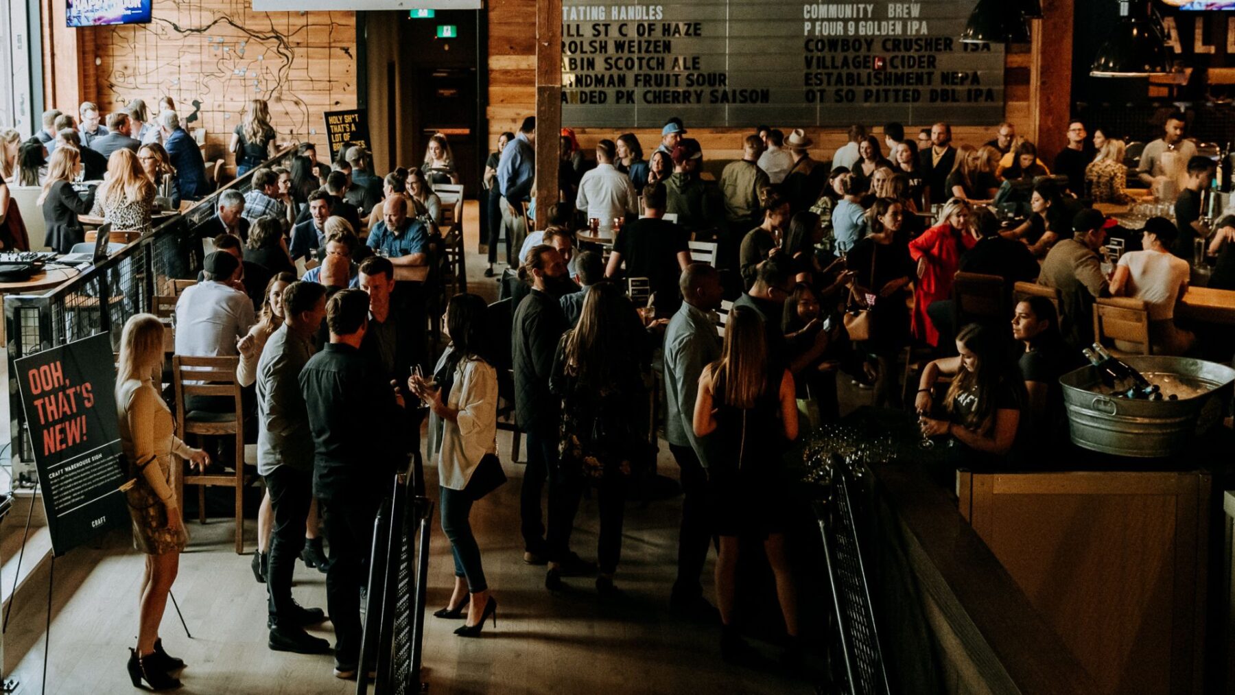 CRAFT Beer Market A large group of people socializing in a busy, modern bar with wooden décor, ambient lighting, and a drink menu featuring craft beers displayed on the wall.