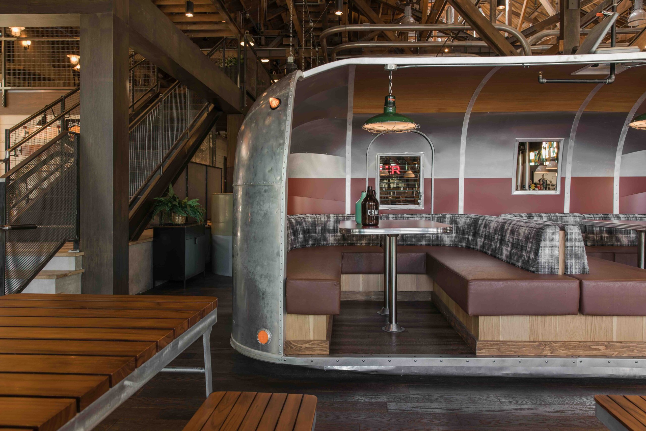 CRAFT Beer Market A restaurant booth designed to look like an Airstream trailer interior, with plaid seating, a pendant lamp, and a wooden table, set in an industrial-style space.