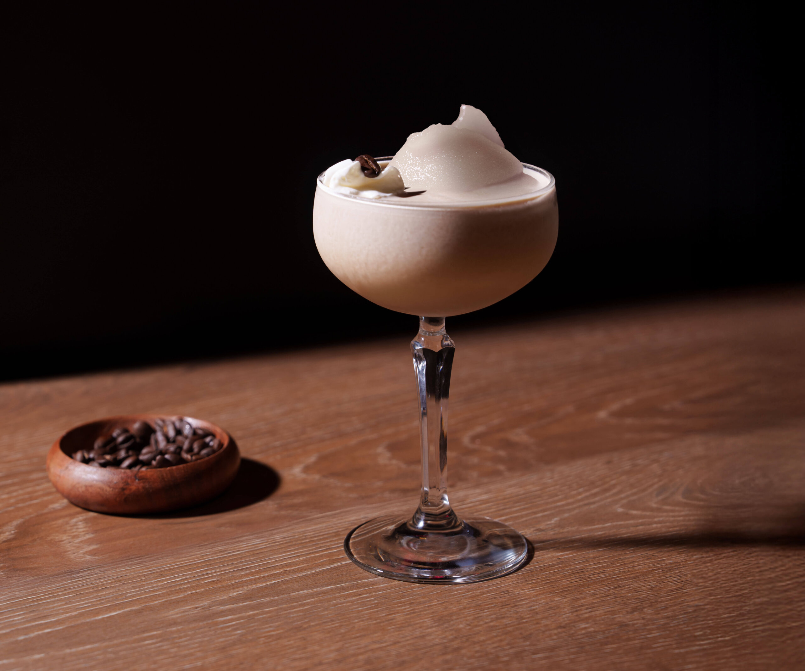 CRAFT Beer Market A creamy craft cocktail served in a coupe glass with a large ice shard, garnished with coffee beans, sits on a wooden table. A small bowl of coffee beans is nearby—perfect for craft cocktail lovers and group bookings.