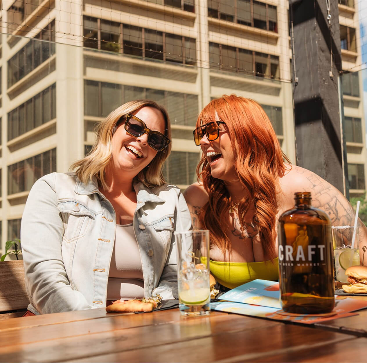 CRAFT Beer Market Two women sit at an outdoor table on a sunny day, smiling and laughing over craft cocktails, with drinks and food in front of them and city buildings in the background.