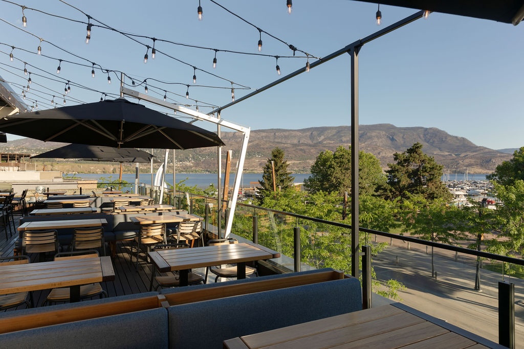 CRAFT Beer Market Outdoor restaurant patio with wooden tables, string lights, and large umbrellas overlooking a lake, marina, and mountains on a clear day.