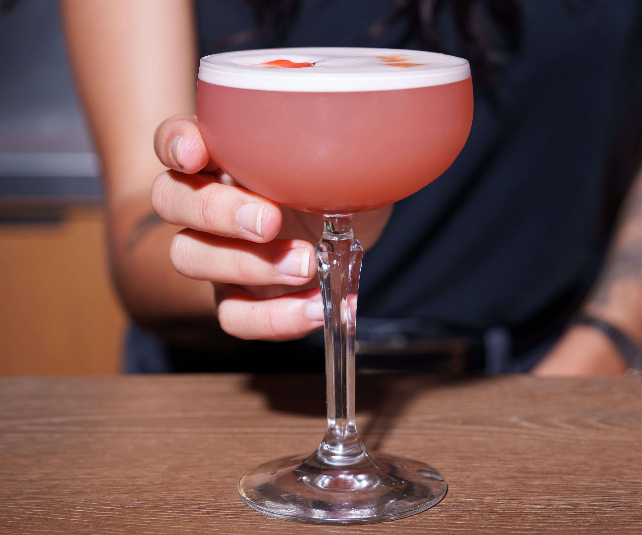 CRAFT Beer Market A person holds a coupe glass filled with a pink craft cocktail topped with foam, set on a wooden surface.