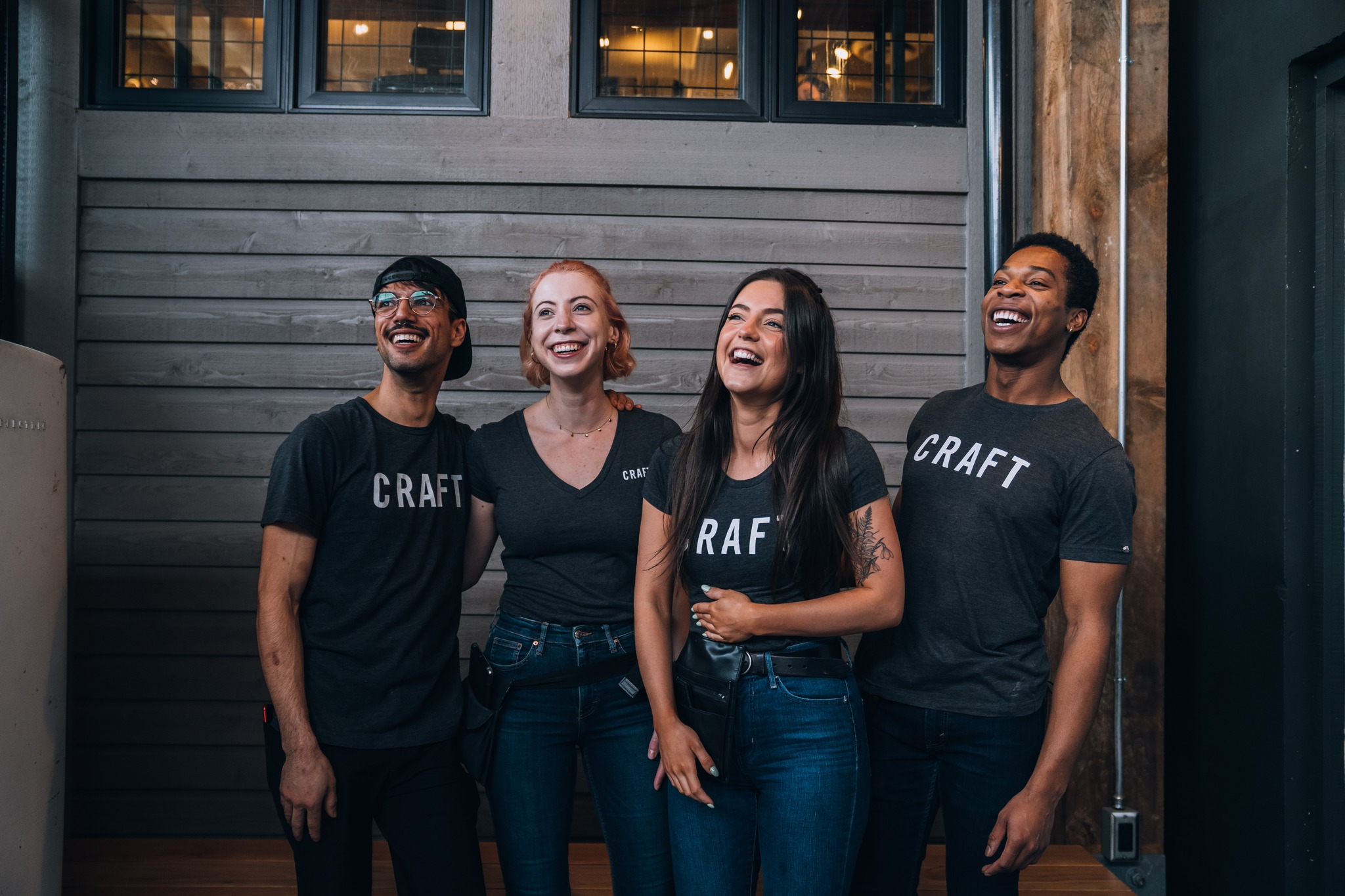CRAFT Beer Market Four people wearing matching "CRAFT" t-shirts stand together indoors, smiling and laughing in front of a wooden wall—capturing the fun spirit of Craft Beers enjoyed at multiple locations.