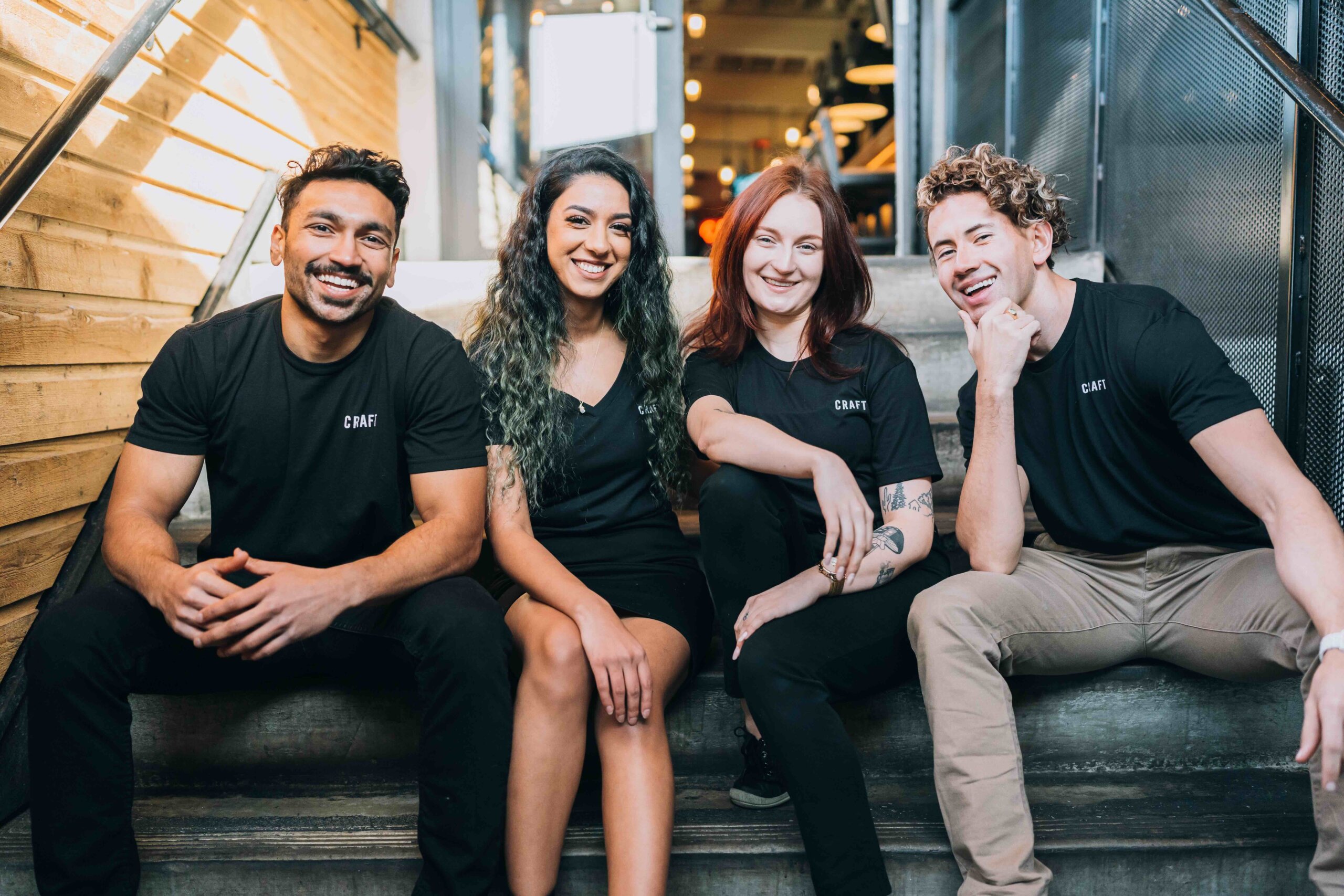 CRAFT Beer Market Four people in matching black shirts sit on indoor stairs, smiling and posing for a group photo.