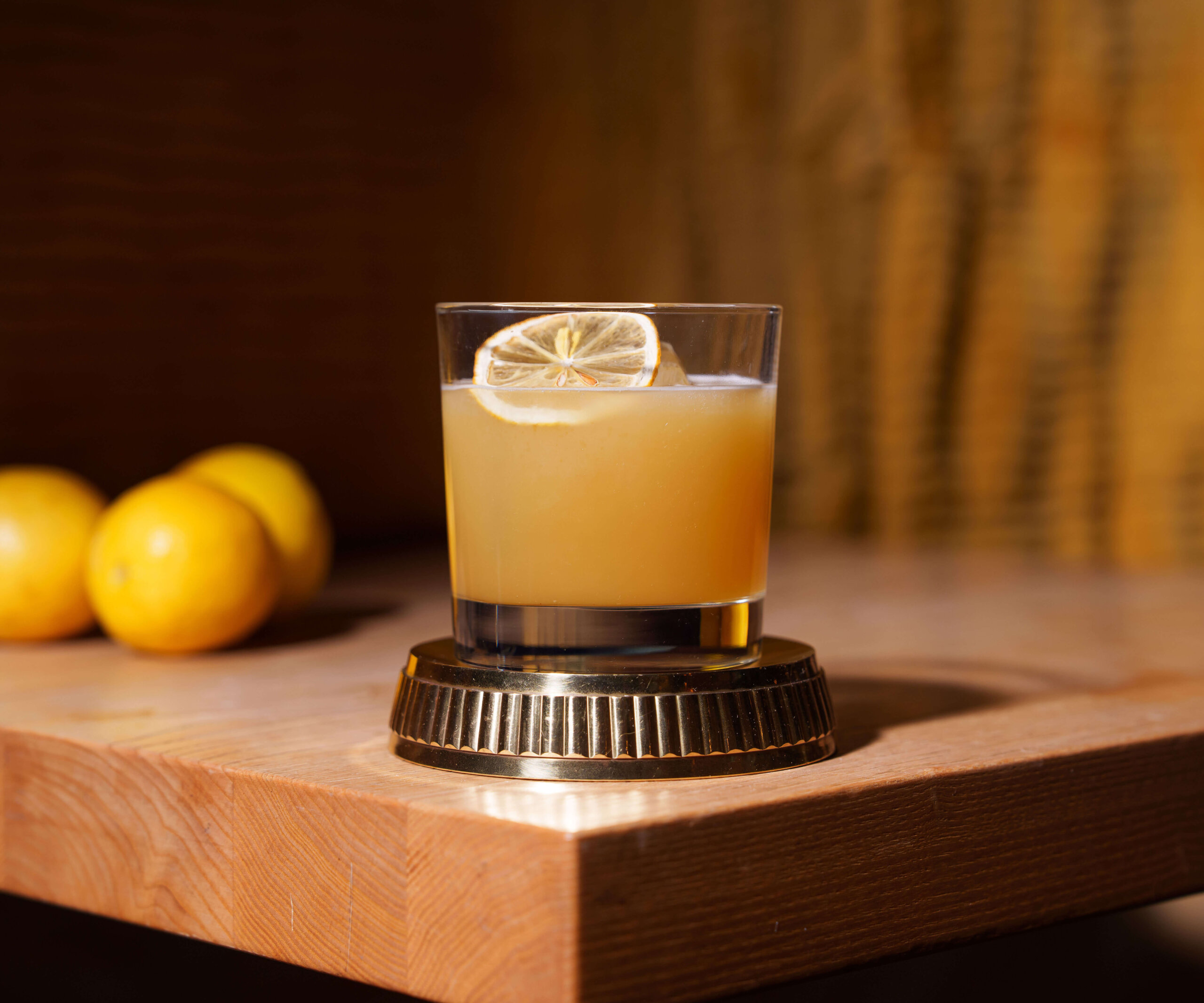 CRAFT Beer Market A glass of orange-brown craft cocktail garnished with a dried lemon slice sits on a brass coaster on a wooden table, with lemons in the background.