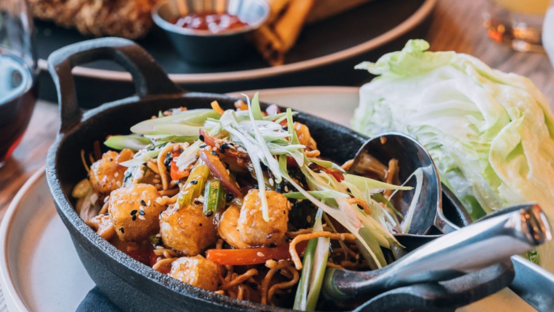CRAFT Beer Market A cast iron skillet with stir-fried noodles, vegetables, tofu, and green onions, served with a side of fresh lettuce—perfect to enjoy alongside craft beers at any of our multiple locations.