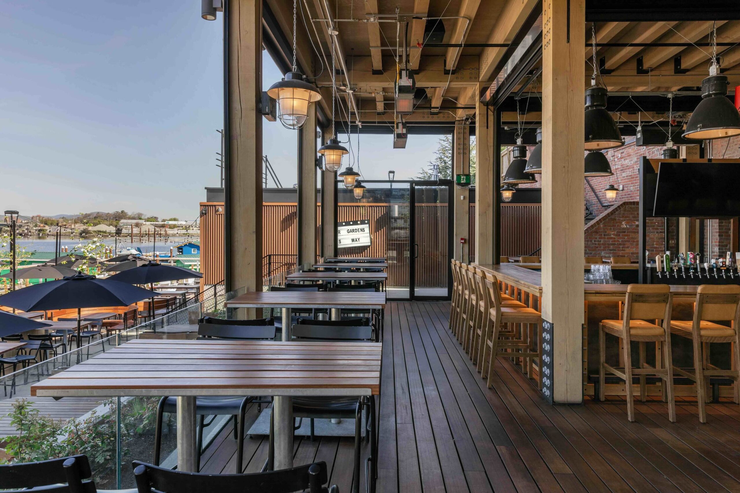 CRAFT Beer Market Outdoor restaurant patio with wooden tables and chairs, high bar seating, hanging lights, and a view of boats and umbrellas by the waterfront—perfect for enjoying craft beer in a casual dining atmosphere.